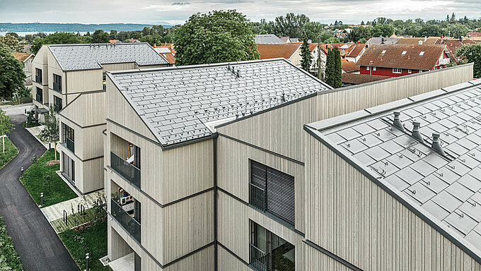 Vue aérienne des immeubles d'appartements modernes à Utting am Ammersee, Allemagne. Le projet résidentiel impressionne par sa construction hybride en bois innovante et les R.16  PREFA durables dans la couleur P.10 gris sombre. Les bâtiments s'intègrent harmonieusement dans l'environnement verdoyant et offrent une combinaison esthétique de façades en bois et de toiture en aluminium de haute qualité. L'installation précise des panneaux de toiture assure la résistance aux intempéries et la durabilité, tandis que les lignes épurées et l'architecture moderne créent un environnement de vie accueillant et durable.