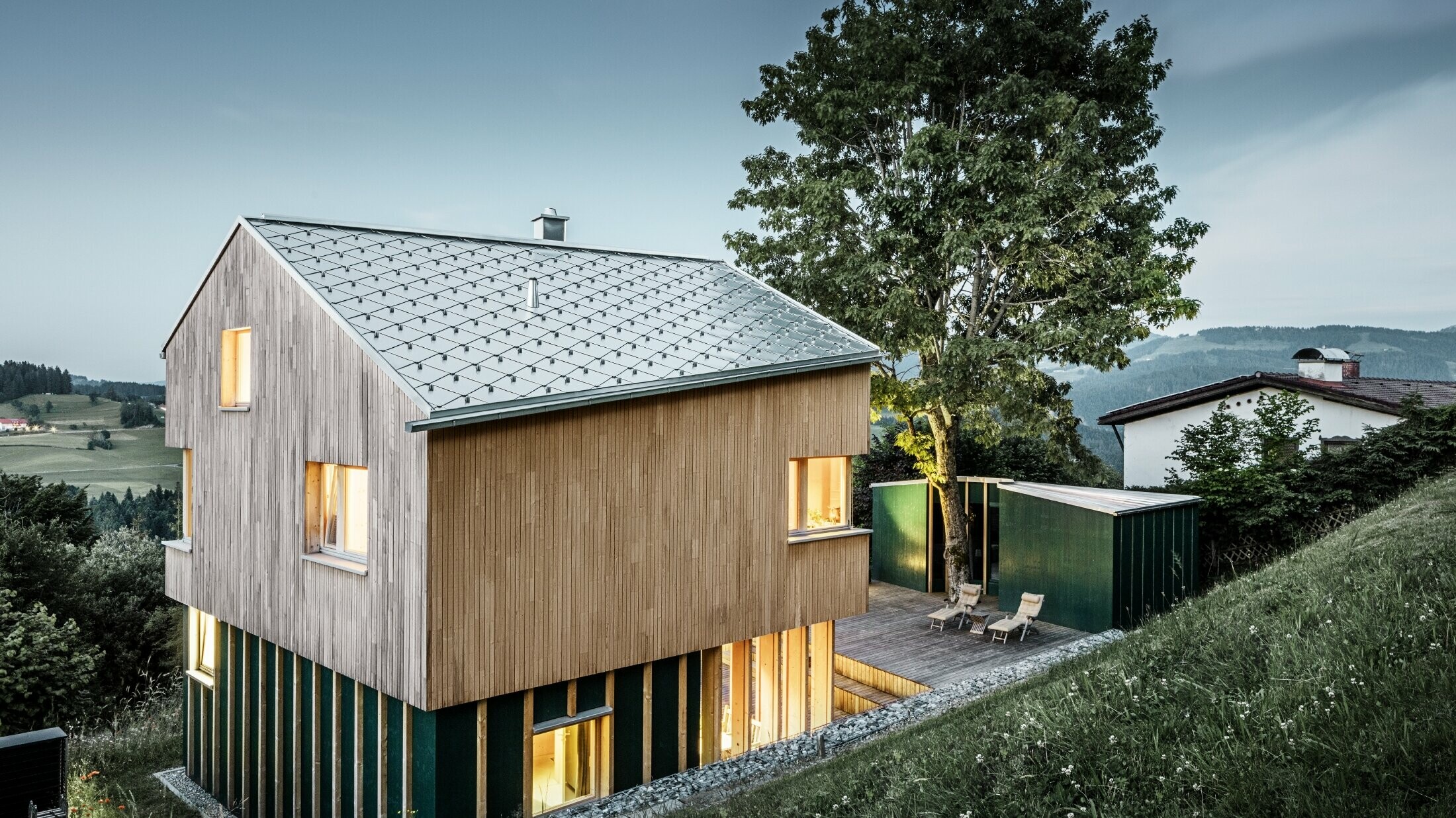 Ein Einfamilienhaus in Hanglage wurde mit der PREFA Dachraute 44x44 in Naturblank eingedeckt. Das langlebige Aluminiumdach harmoniert perfekt mit der hellen Holzfassade. Die Fenster des Gebäudes strahlen Licht und Wärme aus und kontrastieren mit der abendlichen Dämmerung im Hintergrund. 