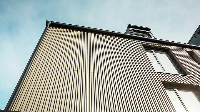 La photo montre la façade d'un immeuble moderne de Feldkirchen, conçu par l'architecte Stefan Heigl. La façade est revêtue de Prefalz de couleur bronze et possède de grandes fenêtres qui laissent entrer beaucoup de lumière à l'intérieur. La gouttière et le toit à pignon sont en gris noir, également avec les matériaux Prefalz. Le design élégant et contemporain allie des matériaux de haute qualité et une architecture fonctionnelle à la fois esthétique et durable.