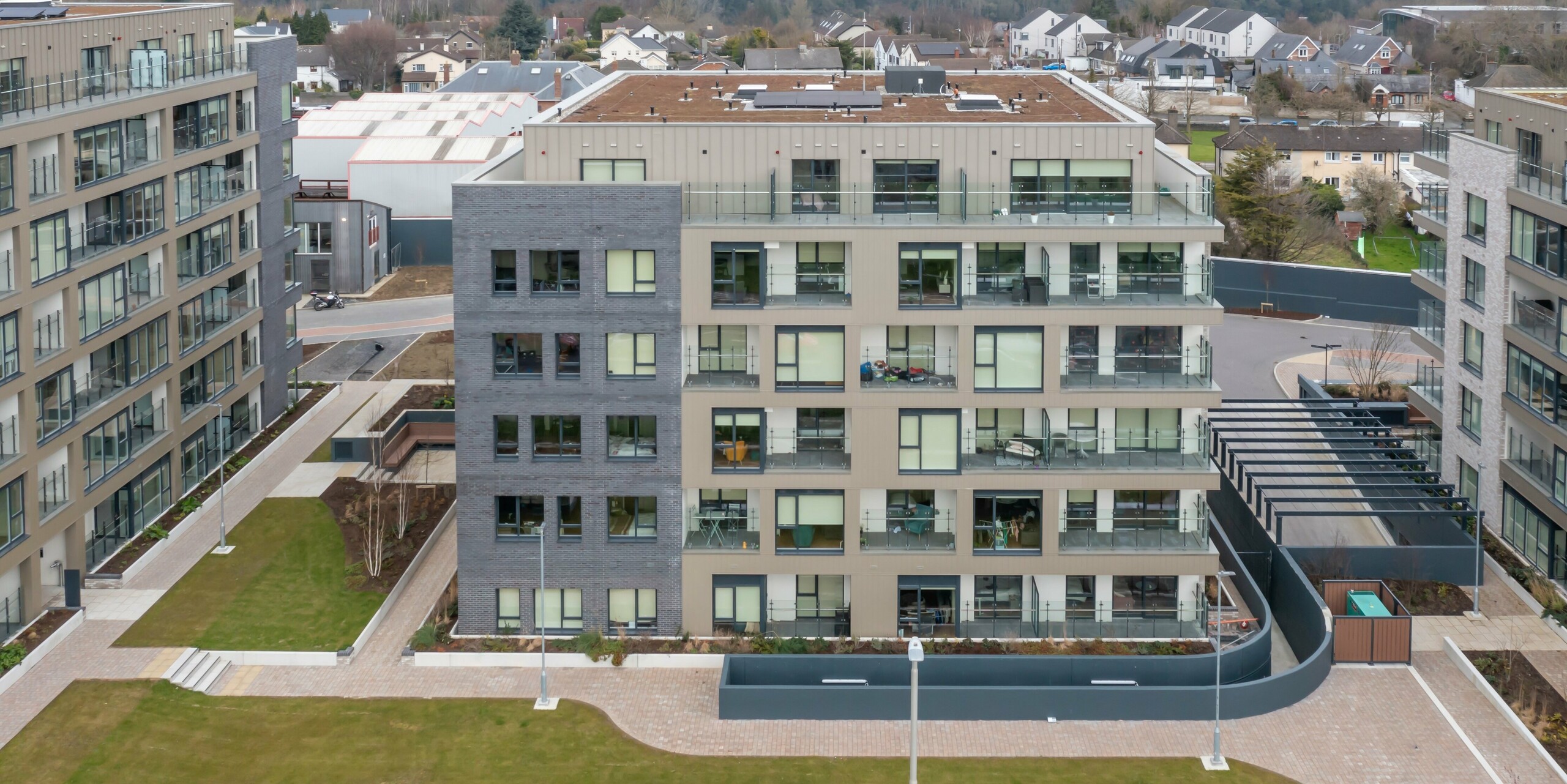 Prise de vue par drone du complexe résidentiel Palmers Gate à Palmerstown, Dublin, Irlande. Le complexe immobilier comprend plusieurs blocs résidentiels avec un revêtement PREFA de couleur bronze et un système de façade PREFALZ. La façade combine un revêtement en aluminium de haute qualité avec des éléments en brique et de grandes fenêtres en verre. Les espaces extérieurs sont aménagés avec des espaces verts, des allées et des jardinières, offrant aux résidents un environnement agréable. Au total, environ 1 700 m² de revêtement PREFA et environ 1 200 m² de système de façade PREFALZ ont été utilisés. Les produits en aluminium sont reconnus pour leur durabilité et leur aspect esthétique.