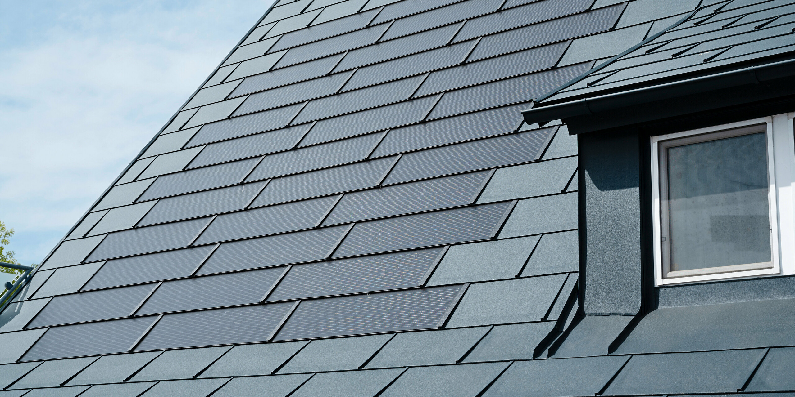 Vue détaillée du toit rénové d'une maison individuelle à Leonding avec des tuiles solaires PREFA de couleur anthracite. Le gros plan montre les panneaux en aluminium de haute qualité de PREFA, qui permettent une intégration transparente des modules solaires. Cette solution durable et résistante aux intempéries offre un moyen durable et économe en énergie de produire de l’électricité. L'image illustre la qualité esthétique et fonctionnelle des matériaux utilisés. Sur le bord droit de l’image, vous pouvez voir une fenêtre blanche sous une lucarne.