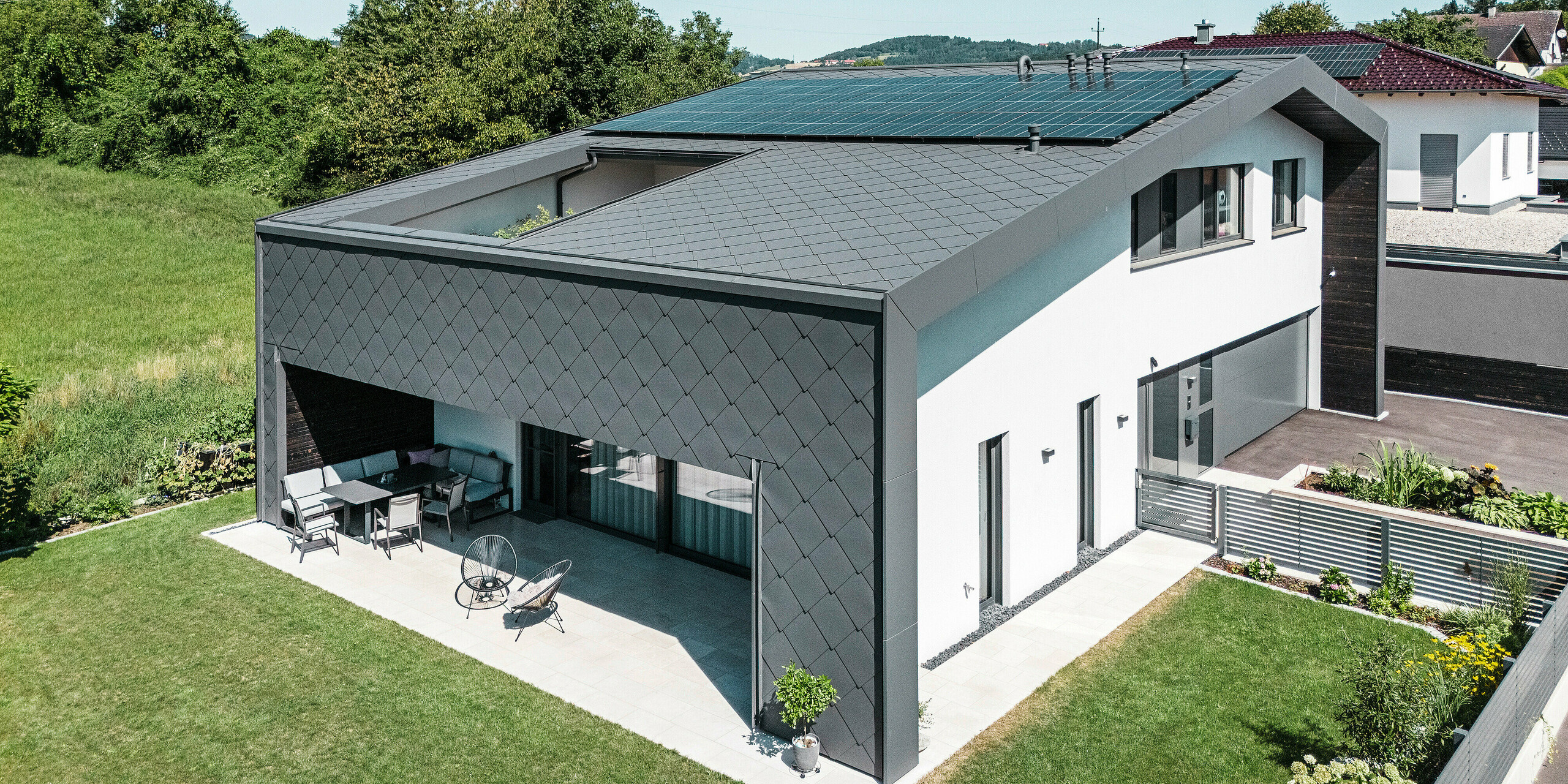 Maison familiale moderne à Aschach avec un élégant revêtement en aluminium en losanges de toiture et de façade 44x44 en revêtement P.10 gris sombre. Le bâtiment se caractérise par une spacieuse terrasse couverte qui s'ouvre harmonieusement sur le jardin bien entretenu. Une installation photovoltaïque sur le toit assure une production d’énergie durable. La combinaison d'une architecture moderne, d'une coque en aluminium résistante aux intempéries et d'une simple façade en plâtre blanc donne à la maison une apparence élégante et contemporaine.