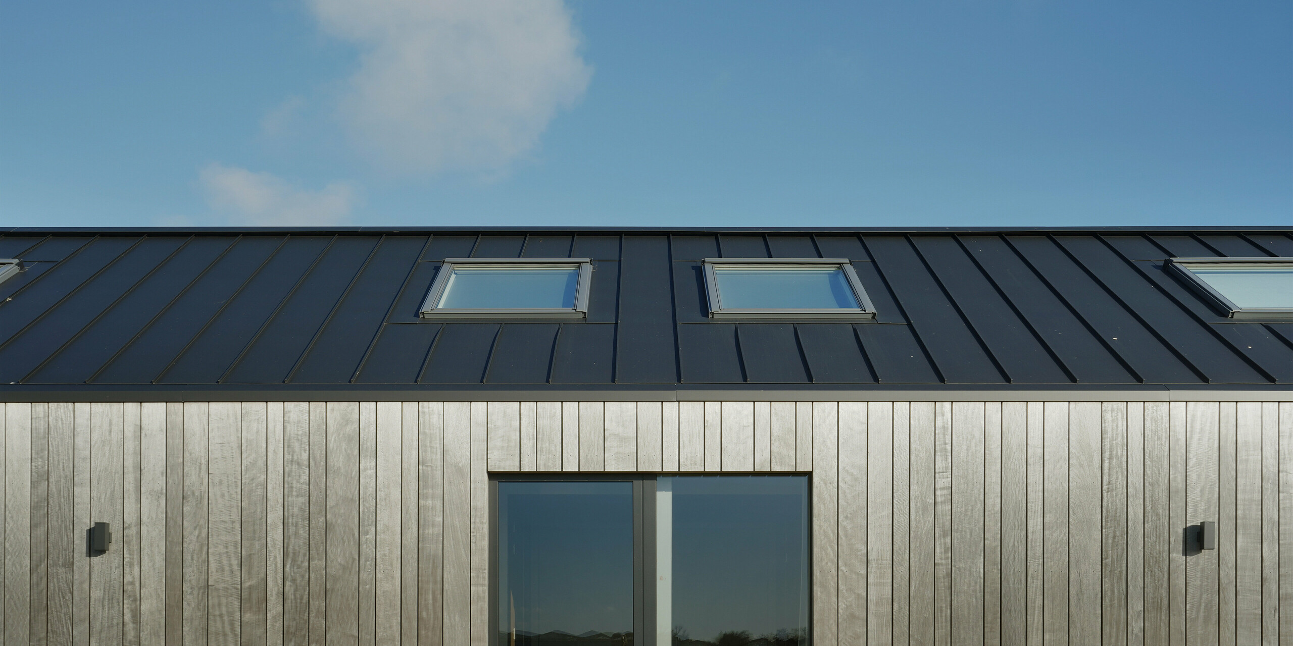 Gros plan sur la toiture à joints debout PREFALZ en noir et gris d'une maison familiale moderne à Uithoorn, aux Pays-Bas. L'image met en évidence l'intégration précise des fenêtres de toit dans le toit en aluminium de haute qualité. La combinaison de matériaux robustes et résistants aux intempéries et d'éléments en bois naturel sur la façade souligne le design durable et contemporain de cette grange vivante.