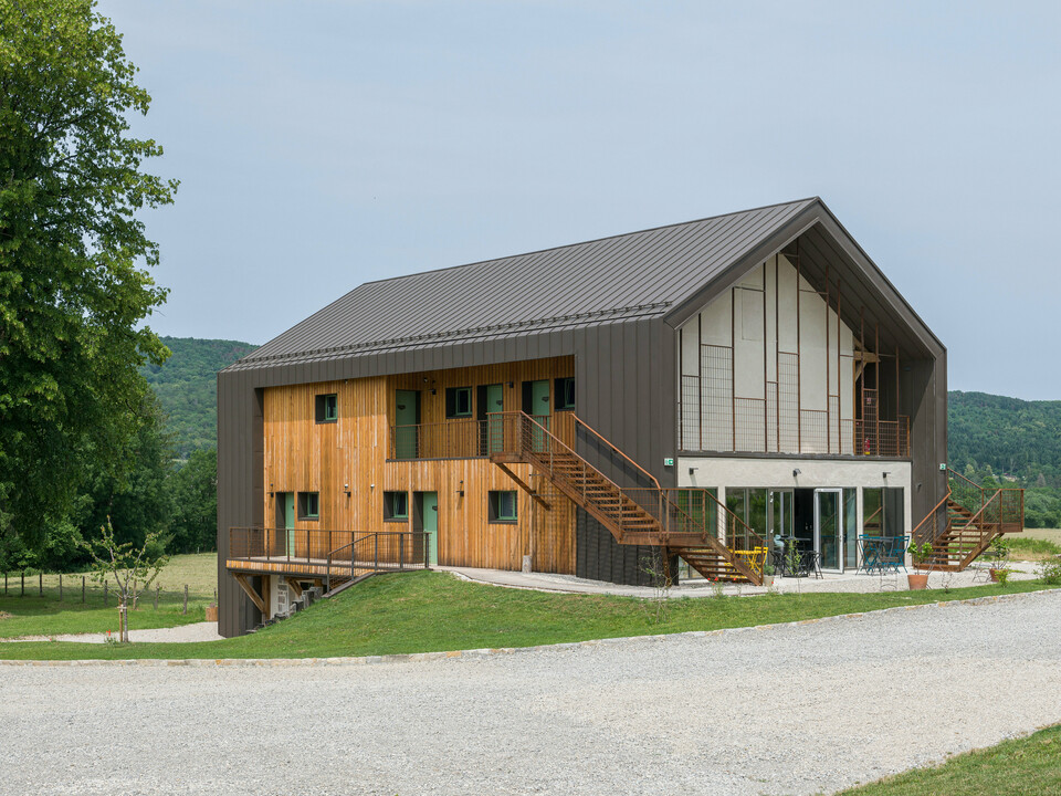 Vue d'angle d'un hôtel contemporain à deux étages avec une coque en aluminium PREFA et une façade en bois naturel. Le toit et certaines parties de la façade ont été revêtus de PREFALZ en brun P.10. Les caractéristiques remarquables du bâtiment sont de grandes fenêtres et un escalier extérieur qui mène à l'étage supérieur. L'Hôtel en Tilleul dispose également d'une terrasse au rez-de-chaussée. Le bâtiment se trouve dans un cadre rural idyllique surplombant des collines. La route d'accès en gravier est visible au bas de la photo.