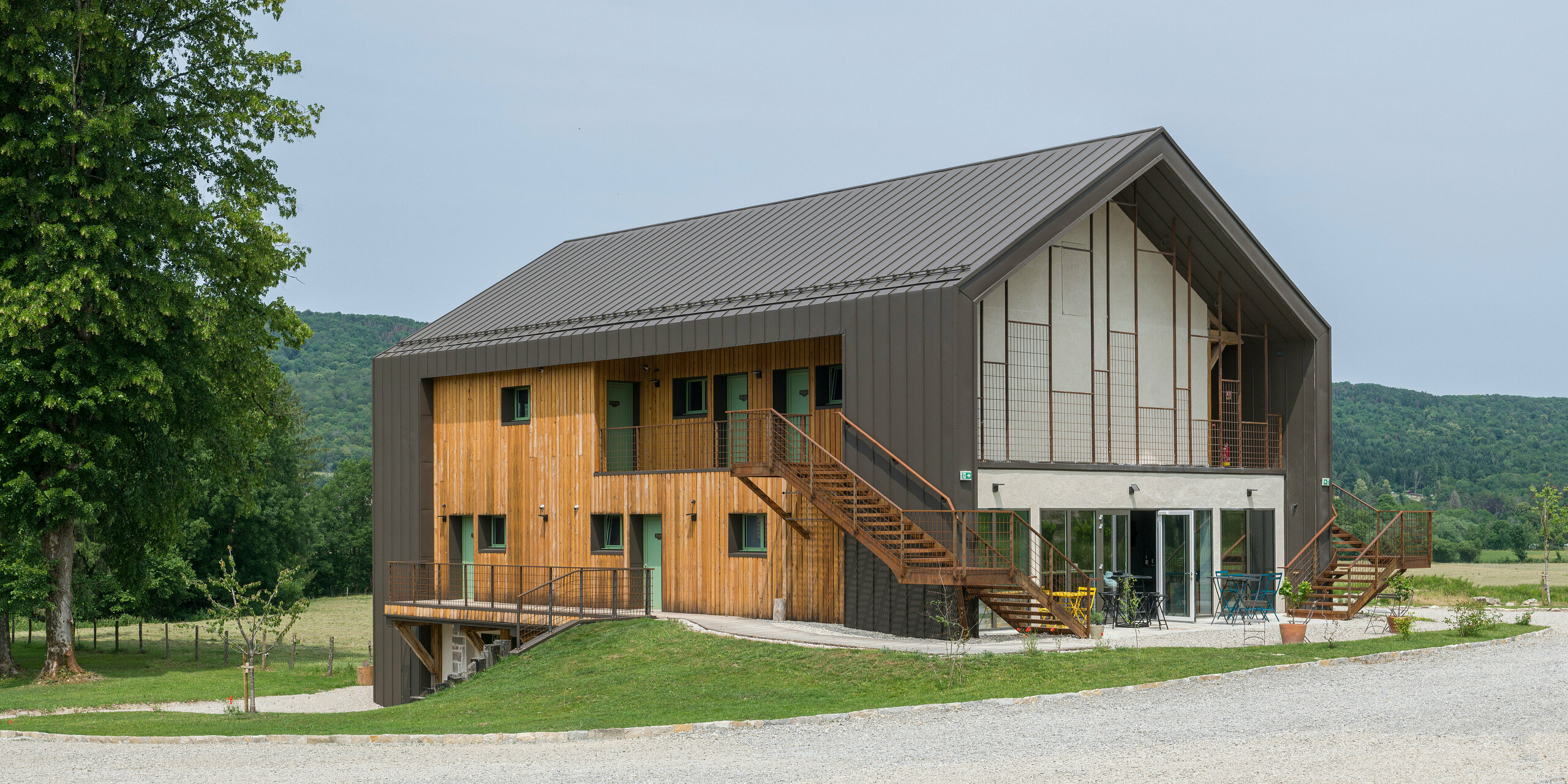 Vue d'angle d'un hôtel contemporain à deux étages avec une coque en aluminium PREFA et une façade en bois naturel. Le toit et certaines parties de la façade ont été revêtus de PREFALZ en brun P.10. Les caractéristiques remarquables du bâtiment sont de grandes fenêtres et un escalier extérieur qui mène à l'étage supérieur. L'Hôtel en Tilleul dispose également d'une terrasse au rez-de-chaussée. Le bâtiment se trouve dans un cadre rural idyllique surplombant des collines. La route d'accès en gravier est visible au bas de la photo.