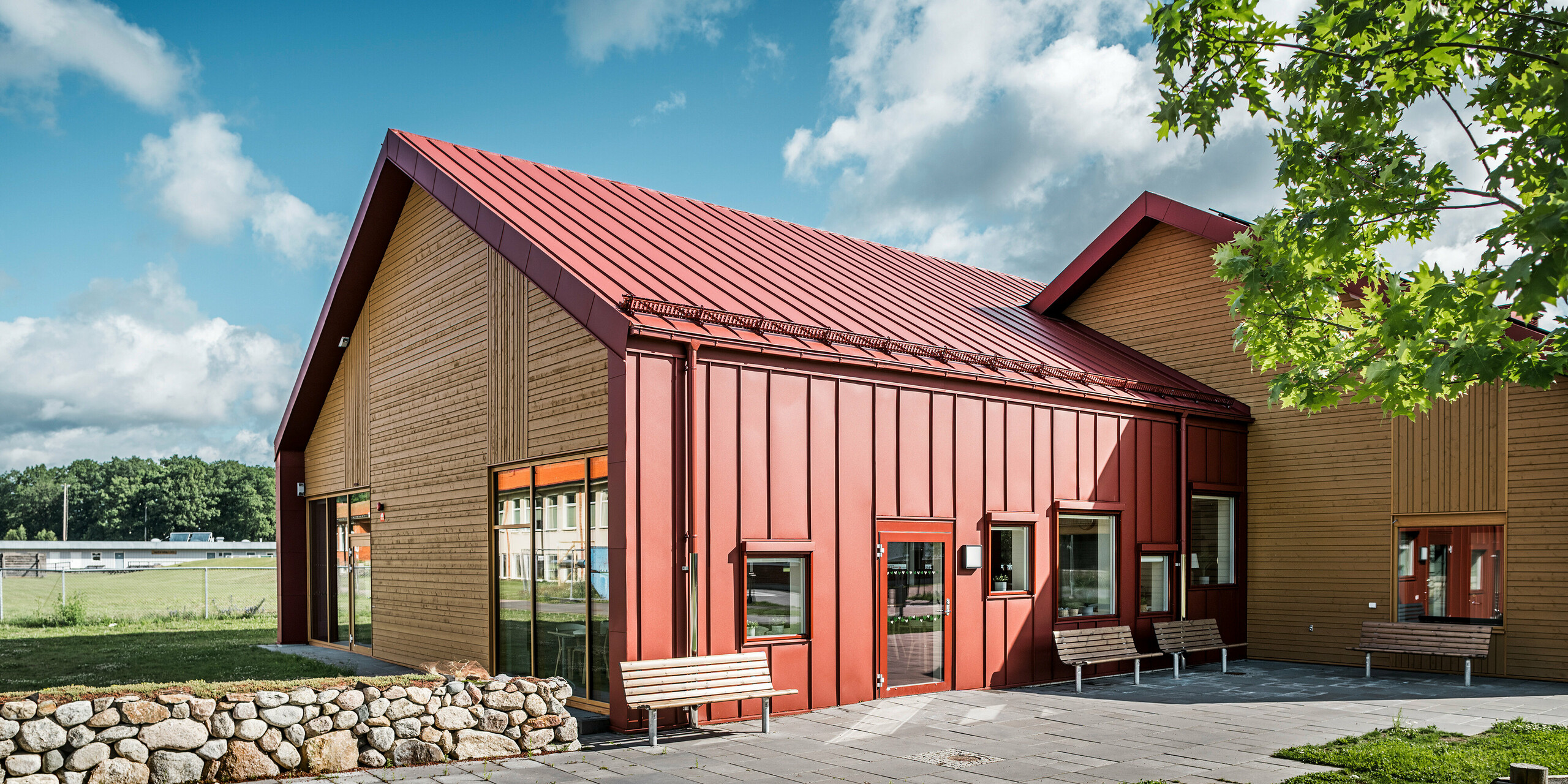 Vue extérieure de la cantine scolaire Restaurang Gröna Hjärtat à Broby, Suède. La photo montre des bancs devant le bâtiment, qui présentent une combinaison frappante de PREFALZ rouge oxyde et de bois au pignon. La conception architecturale met l'accent sur les lignes claires et le contraste harmonieux entre les surfaces en aluminium rouge et le ton chaud du bois. Des murs en pierre et des espaces verts sont visibles au premier plan, encadrant le design moderne du bâtiment. Les environs et le bâtiment se fondent dans un espace accueillant et bien pensé.