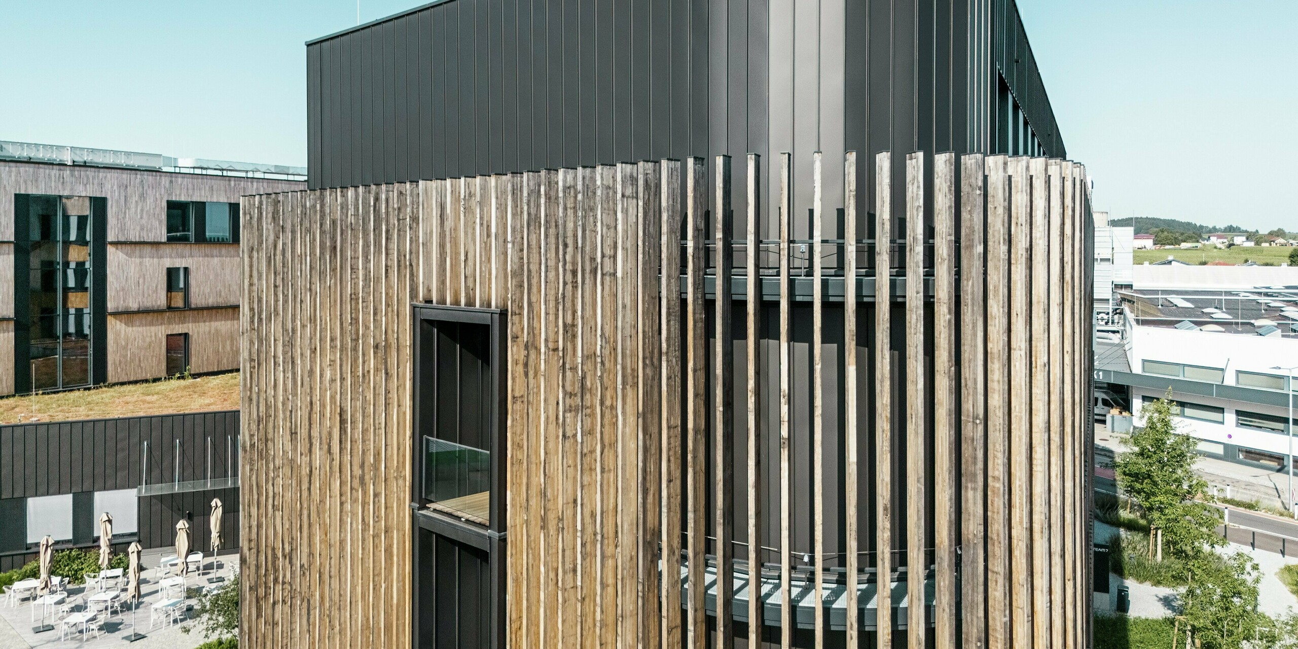 Le monde Team 7 à Ried im Innkreis impressionne par sa façade moderne PREFALZ dans la couleur spéciale noir-brun, associée à des éléments en bois marquants. Le traitement précis de l'aluminium et du bois garantit une interaction harmonieuse entre durabilité et esthétique naturelle. Le revêtement en aluminium résistant aux intempéries garantit la durabilité, tandis que le design confère un caractère architectural distinctif. Le ciel bleu brille au-dessus du grenier.