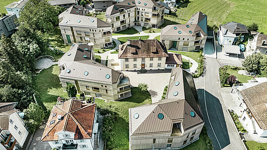 Vue aérienne du complexe résidentiel moderne de Liebwylen, en Suisse. Les bâtiments sont conçus avec des toits en aluminium PREFALZ de couleur brune et disposent de fenêtres de toit rondes. Au centre se trouve la villa historique, entourée de nouveaux bâtiments harmonieusement disposés, le tout intégré dans un paysage verdoyant.