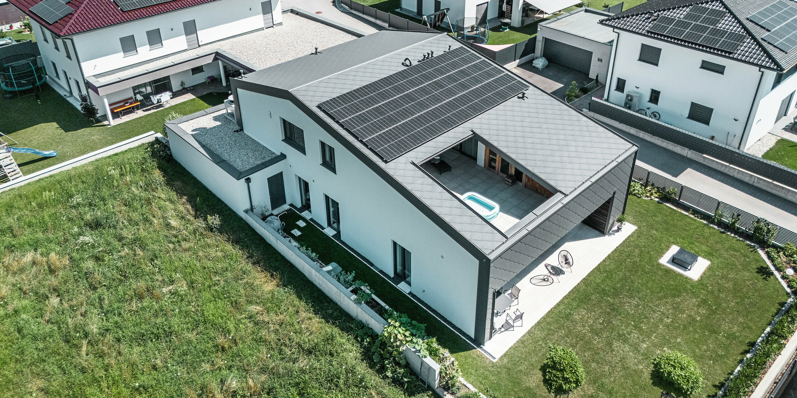 Prise de vue par drone de l'arrière d'une maison familiale moderne à Aschach avec un grand jardin. La façade et le toit sont recouverts de losanges de toiture et de façade 44x44. La surface du toit est équipée d'un système photovoltaïque et d'un atrium qui offre un espace de vie extérieur supplémentaire. La terrasse couverte au rez-de-chaussée et l'atrium confèrent à la maison une atmosphère ouverte et lumineuse. La combinaison d'un revêtement en aluminium et d'une façade en plâtre blanc crée une apparence générale harmonieuse et moderne.