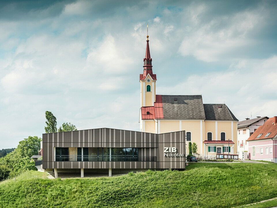 Le centre événementiel ZIB Nikolai à St. Veit dans le sud de la Styrie avec une façade en aluminium moderne et dynamique dans des tons gris brun-beige, au premier plan une prairie verte, à l'arrière-plan l'église Saint-Nicolas avec une façade jaune et un toit rouge.