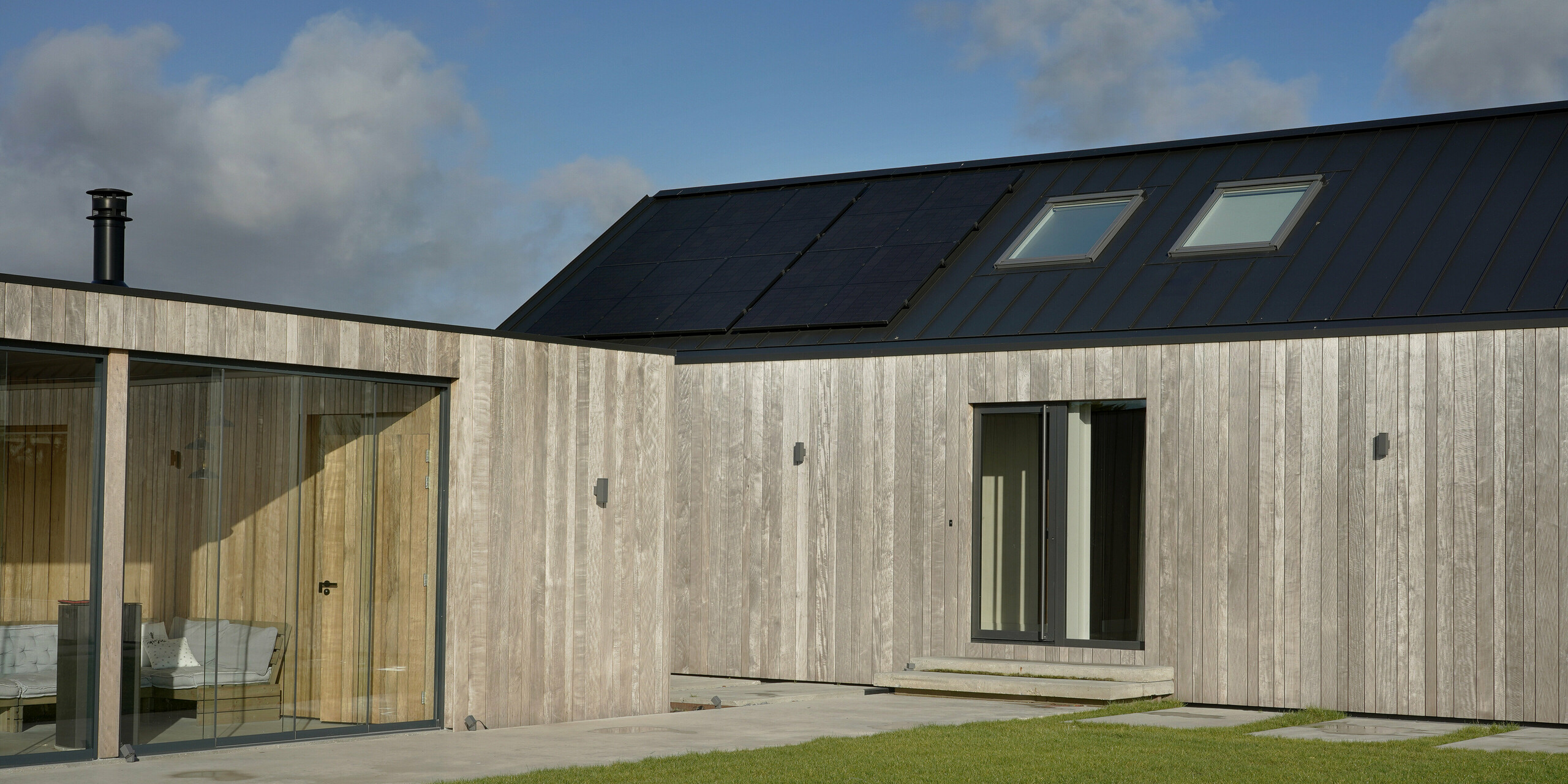 Vue détaillée d'une maison familiale moderne à Uithoorn, aux Pays-Bas, avec toiture à joints debout PREFALZ en gris noir. Le bâtiment présente une combinaison de grandes façades en verre et d'éléments en bois durables. Le toit à pignon est équipé d'aluminium de haute qualité de PREFA, qui garantit durabilité et résistance aux intempéries. L'image met l'accent sur l'architecture contemporaine et le design durable.