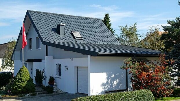 Maison rénovée avec toit à deux pans et garage accolé. Le toit est recouvert de tuiles PREFA et le garage à l’aide de Prefalz couleur anthracite. Un mât avec le drapeau suisse se trouve devant la maison.