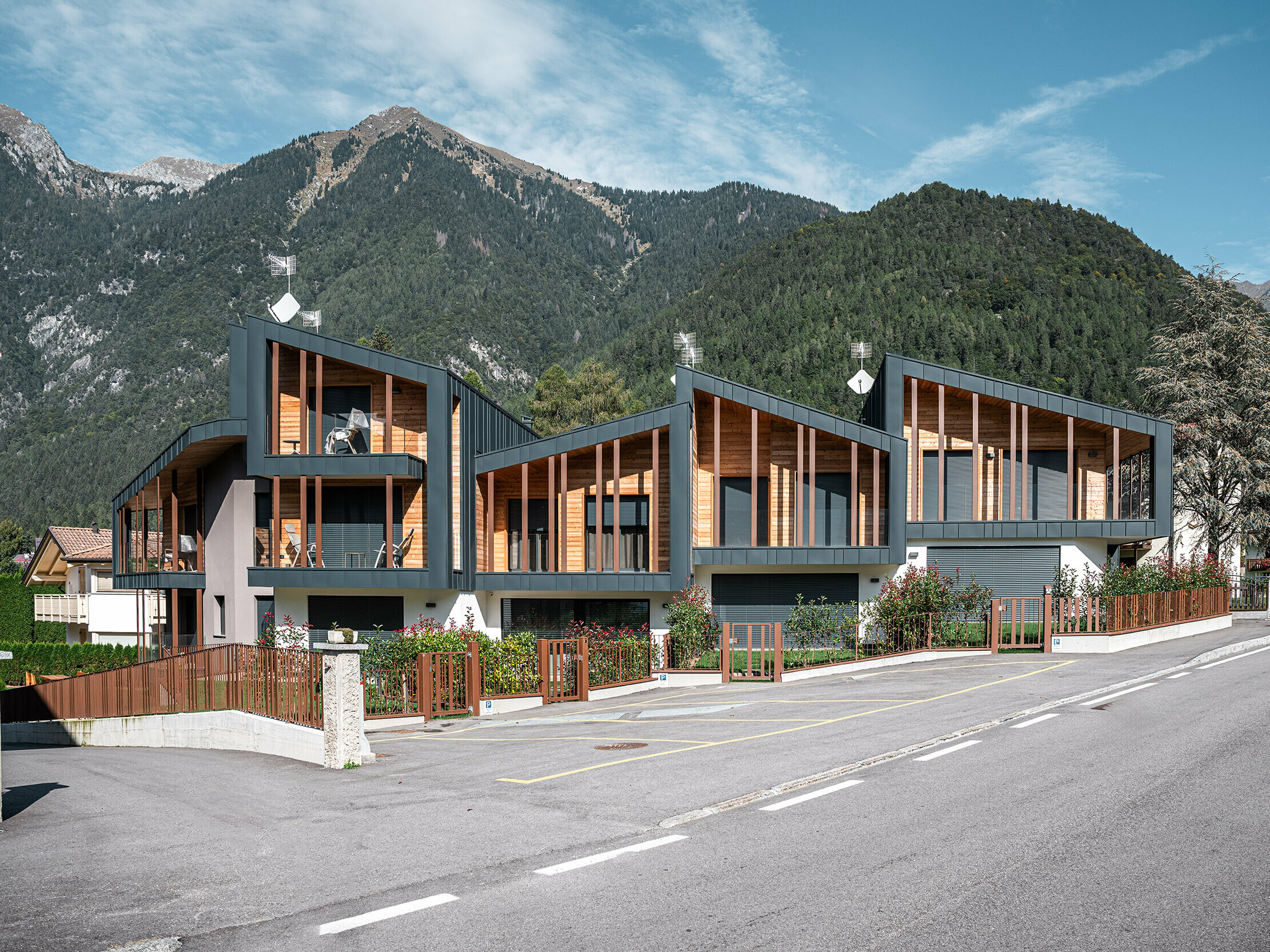 Chalets de vacances modernes avec toits en métal Prefalz couleur anthracite et éléments en bois dans une vallée des Alpes, le long d’une route, avec des jardins clôturés et une vue impressionnante sur les montagnes en arrière-plan.