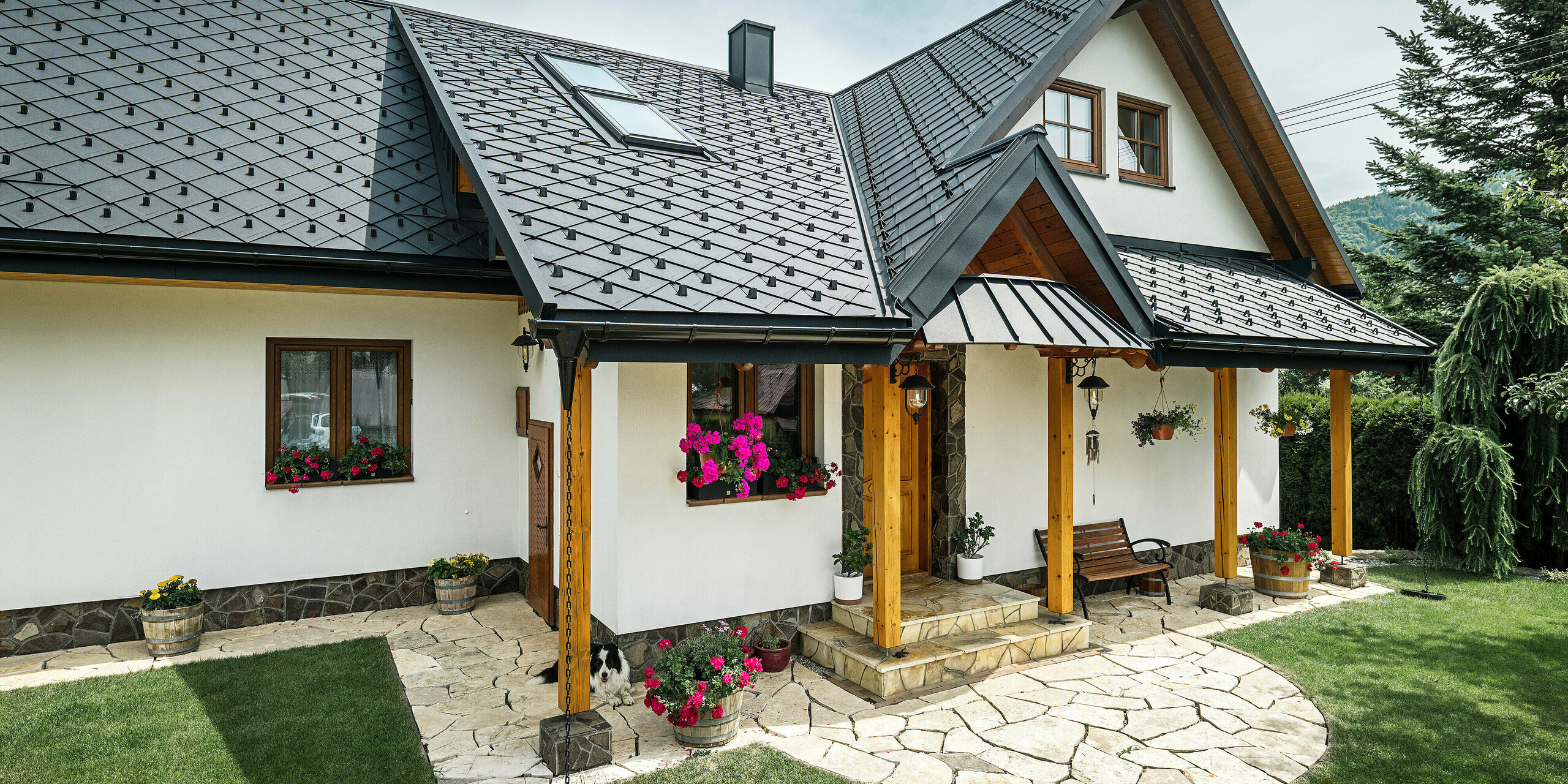 Zone d'entrée d'une maison familiale traditionnelle à Stara Bystrica, Slovaquie, avec un toit en losanges PREFA 29 × 29 en P.10 noir. Le toit en aluminium comprend des fenêtres de toit inclinées et un auvent recouvert de PREFALZ. La zone d'entrée est accentuée par des supports en bois et une terrasse en pierre naturelle. Des compositions florales colorées et un espace vert bien entretenu confèrent à la maison une apparence accueillante et charmante. Le border collie couché devant la maison garde la porte d'entrée. Le chien au repos perfectionne l’image de la vie familiale conventionnelle en milieu rural.