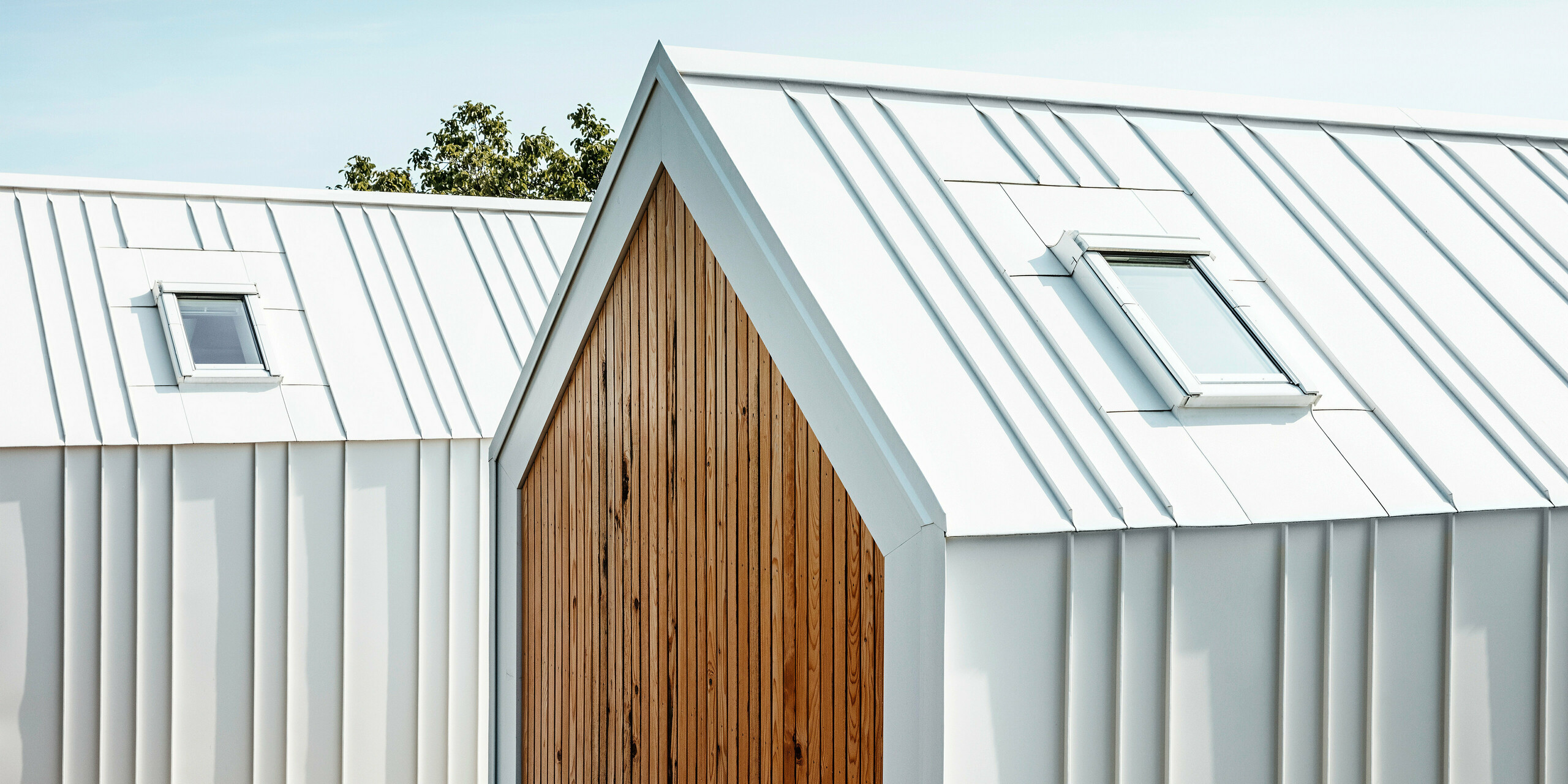 Vue détaillée revêtement de toiture et du de façade PREFALZ blanc des petites maisons « Pri Momi » à Kančevci, Slovénie. Les bâtiments sont conçus dans une combinaison moderne d'aluminium de haute qualité en blanc pur P.10 et de bois naturel. La photo montre les plis exécutés avec précision et la transition élégante du toit à la façade, qui a été réalisée sans aucune interruption visible des joints verticaux. Le choix harmonieux des matériaux souligne le design minimaliste de l'hébergement exclusif.