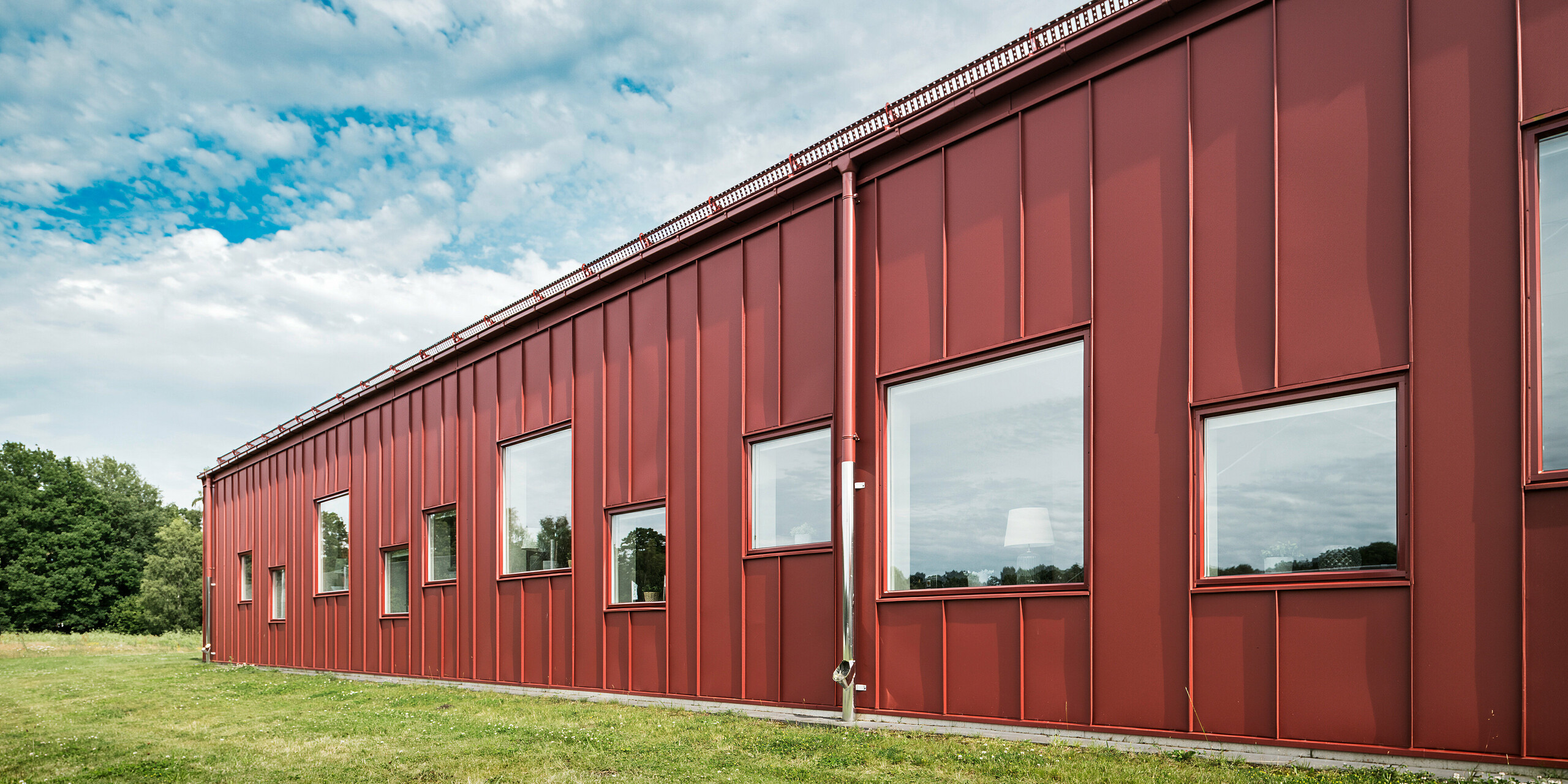 L'arrière de la cantine scolaire Gröna Hjärtat à Broby, en Suède, présente une façade à la fois moderne et robuste en PREFALZ en rouge oxyde. Le revêtement en aluminium durable souligne la haute qualité et la durabilité de la construction. Le système de gouttière est également fini en rouge oxyde, ce qui renforce l'aspect uniforme et souligne l'harmonie esthétique de l'architecture.