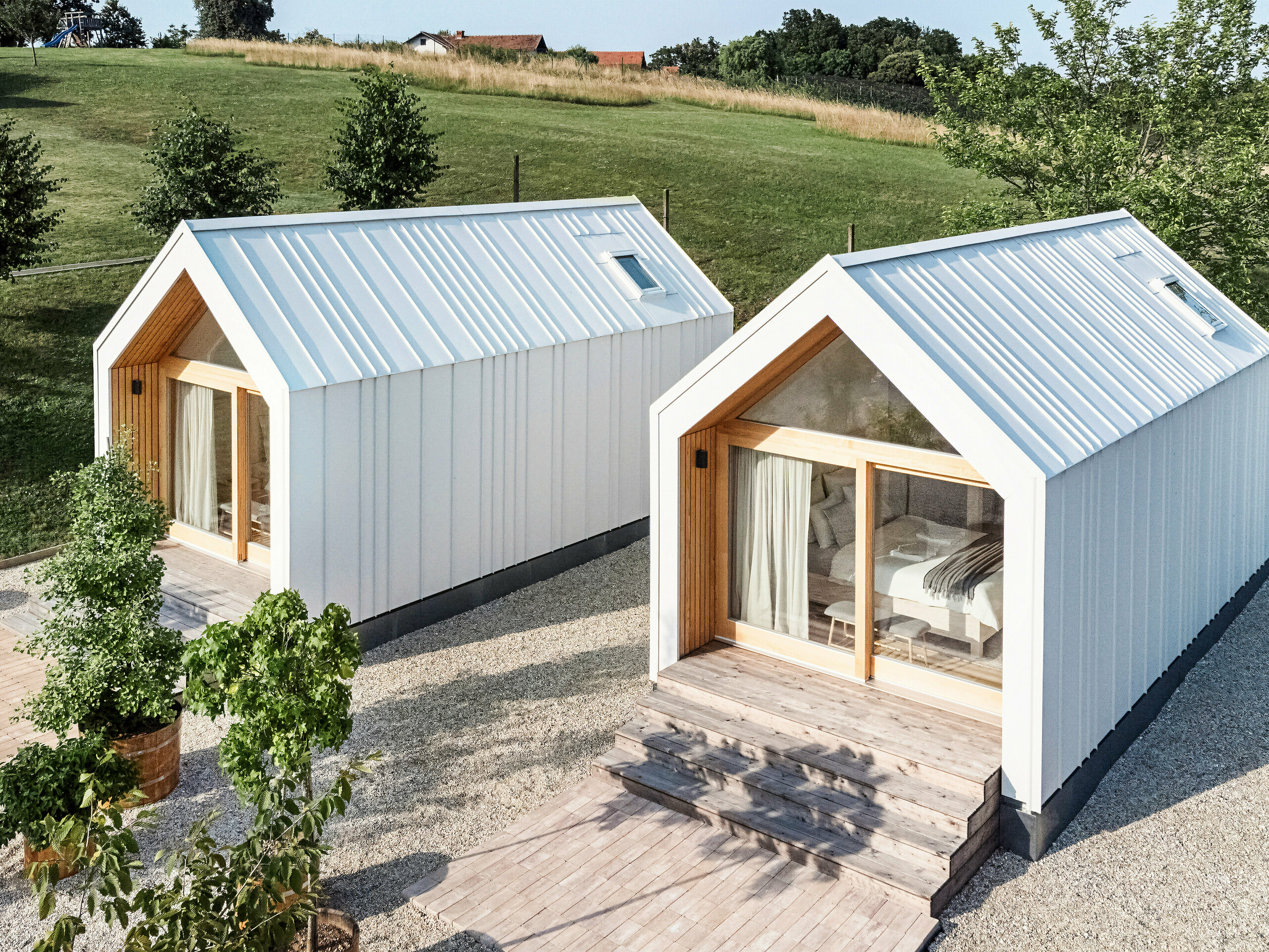 Vue de la façade des deux petites maisons du complexe de vacances « Pri Momi » à Kančevci, en Slovénie. Les micro-maisons ont été construites à ossature bois et dotées d'un revêtement de toit et de façade PREFALZ robuste en blanc pur P.10. Les bâtiments modernes et minimalistes combinent une coque en aluminium à la fois durable et esthétique avec de grandes façades en verre et en bois naturel, intégrés dans un paysage verdoyant. Les spacieuses marches en bois mènent aux entrées, créant une atmosphère chaleureuse et accueillante.