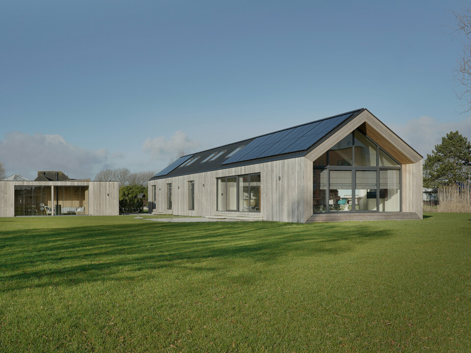 Prise de vue grand angle d'une maison familiale moderne à Uithoorn, aux Pays-Bas, avec une toiture à joints debout PREFALZ en gris noir et un système photovoltaïque intégré. La photo montre l'ensemble de la propriété avec un jardin spacieux et une combinaison de grandes façades vitrées et d'éléments en bois naturel sur la façade. Les lignes claires et les matériaux de haute qualité résistants aux intempéries soulignent la durabilité et le design moderne de la maison. Le bungalow d'un étage a été construit pour répondre aux besoins de vie d'une grande famille.