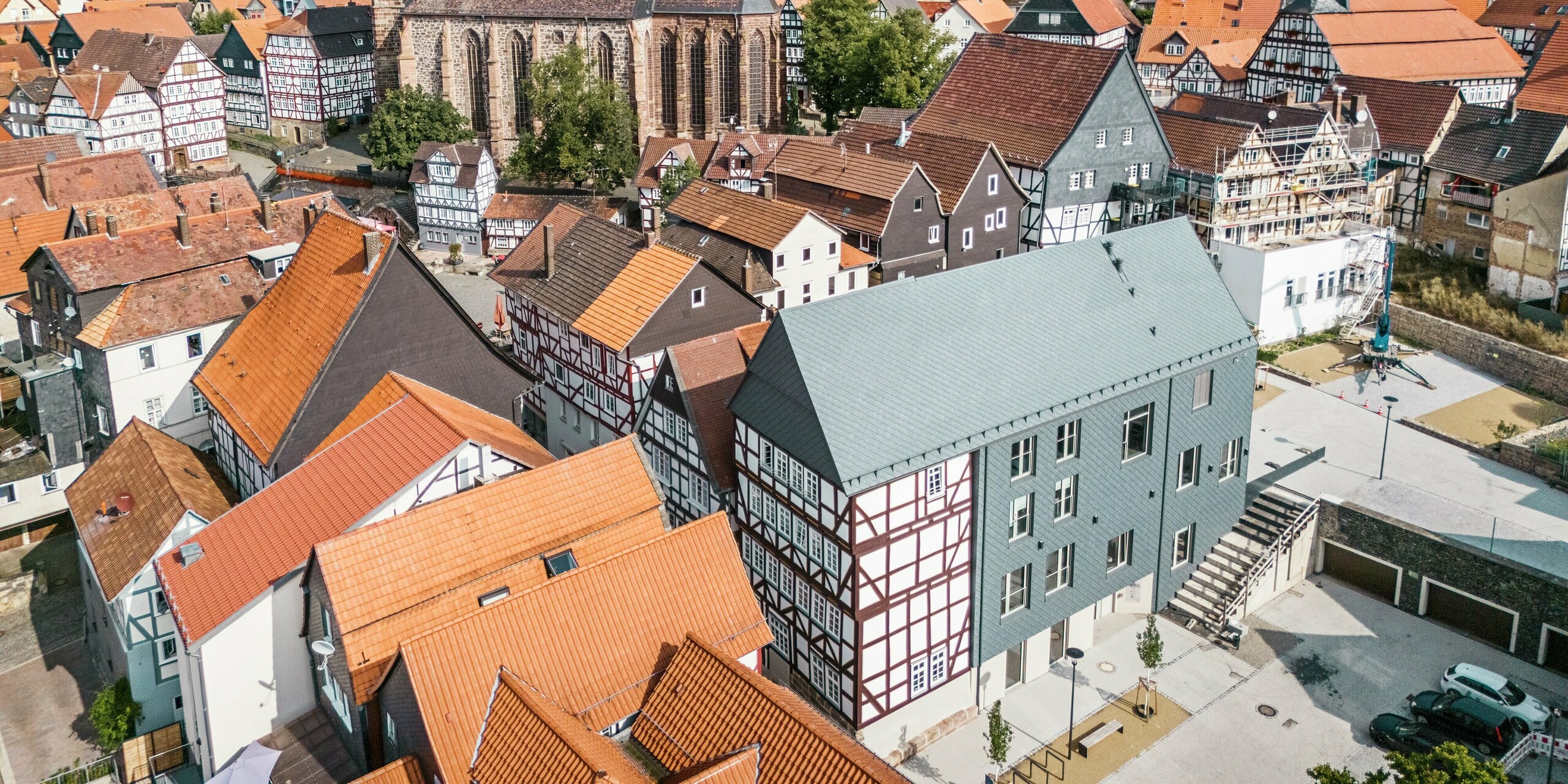 Prise de vue grand angle par drone du Volkshaus à Homberg, DE, avec des losanges de toiture et de façade 29x29 PREFA en P.10 gris pierre. Le design moderne s'harmonise avec la vieille ville historique, caractérisée par des maisons à colombages, une église et un château. Le revêtement en aluminium en forme d'écailles assure durabilité et résistance aux intempéries.