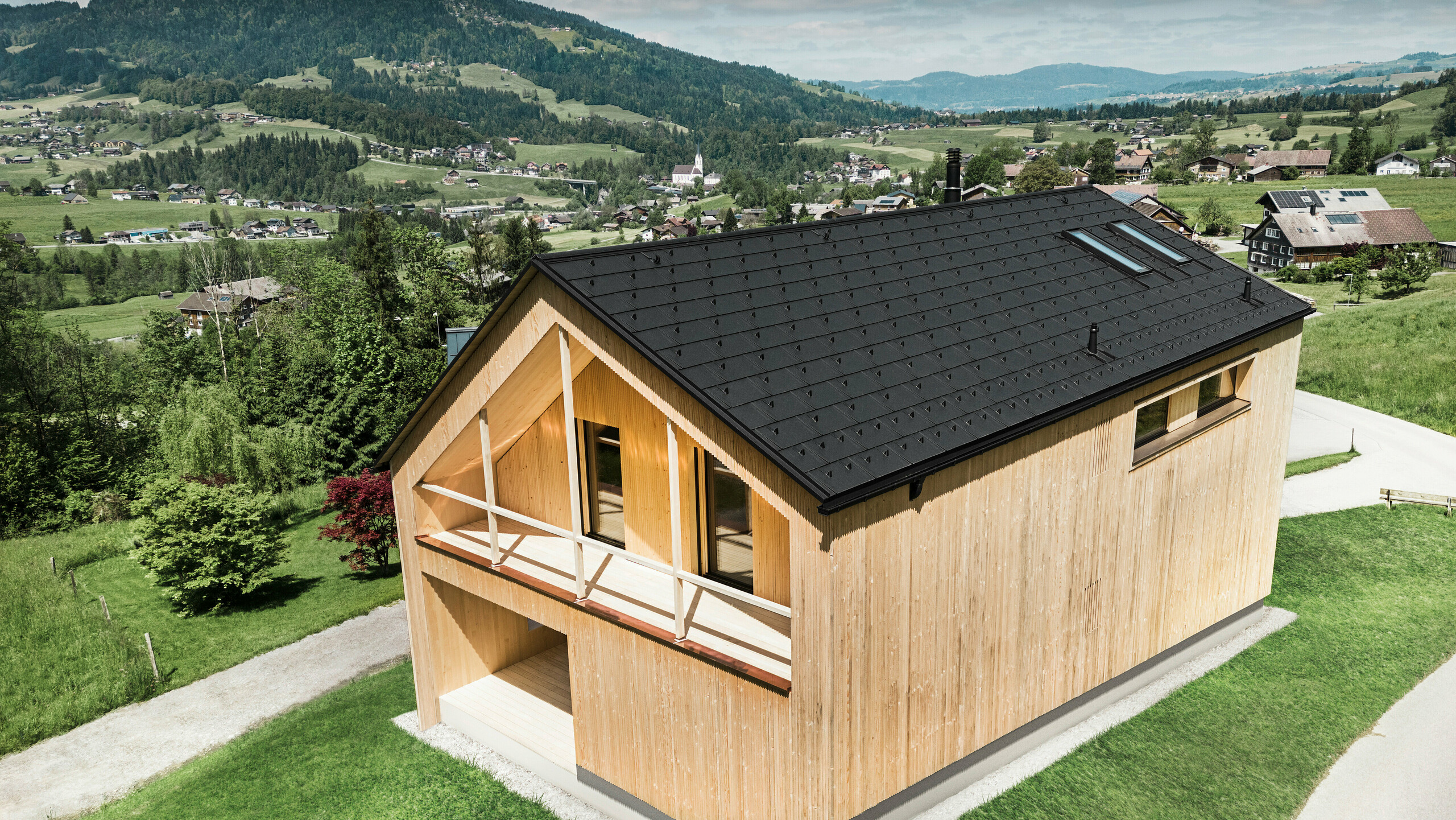 Cette photo montre une maison vitrine écologique à Egg, qui se distingue par sa tuile solaire PREFA et ses R.16 noirs dans le décor pittoresque d'un village autrichien. Les éléments solaires sont parfaitement intégrés dans la structure du toit et fournissent de l'énergie à partir d'une source d'énergie renouvelable, tandis que le bois naturel de la façade et l'environnement verdoyant représentent une combinaison de durabilité et de localité. La vue sur la structure du village environnant avec les bâtiments traditionnels en arrière-plan donne à l'image une dimension supplémentaire et souligne l'intégration réussie de méthodes de construction modernes et respectueuses de l'environnement dans les paysages établis.