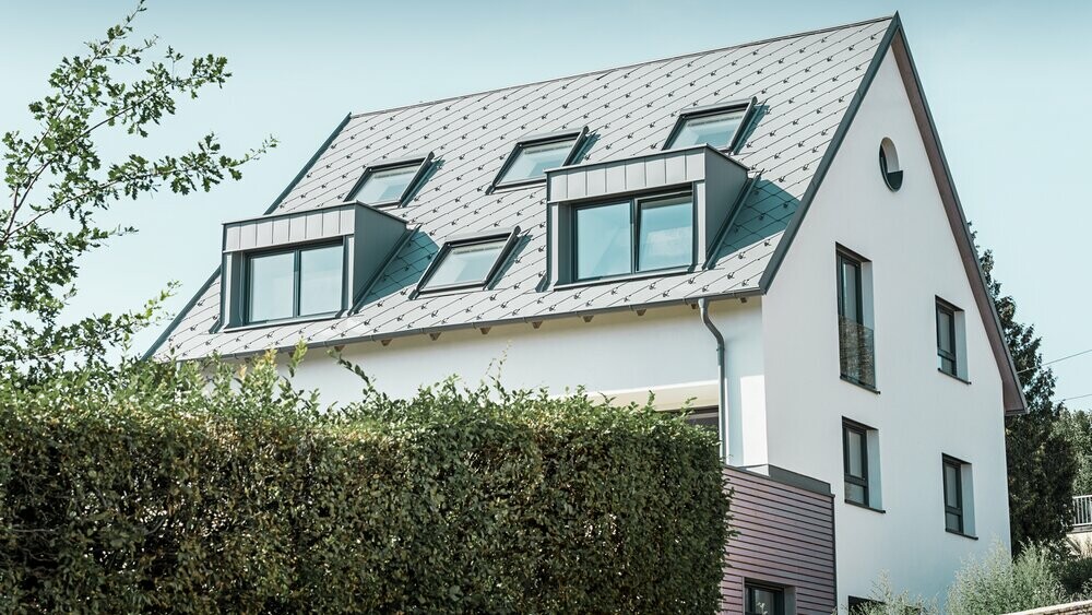 Rénovation d’un toit à deux pans avec deux lucarnes et quatre fenêtres de toit ; la toiture a été recouverte de losanges de toiture 44 × 44 PREFA couleur gris souris. Le choix s’est porté sur une gouttière suspendue PREFA également couleur gris souris. 