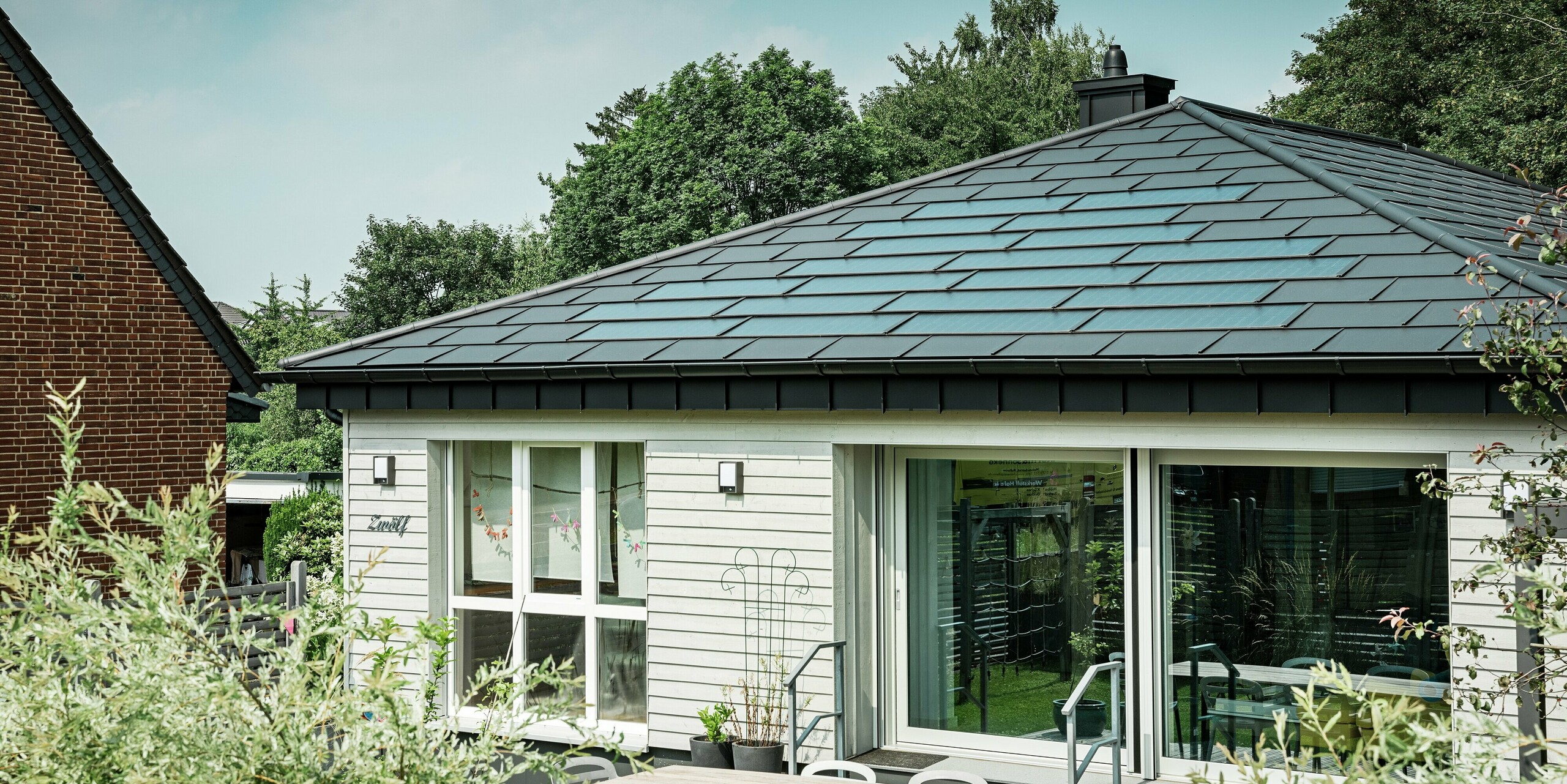 Bungalow rénové à Rommerskirchen avec un élégant toit en aluminium PREFA. Les tuiles solaires PREFA et les R.16 en P.10 noir offrent une protection durable et produisent de l'électricité durable à partir de l'énergie solaire. La généreuse façade vitrée assure une luminosité abondante et le drainage du toit avec gouttière et tuyau d'évacuation allie parfaitement fonctionnalité et design.