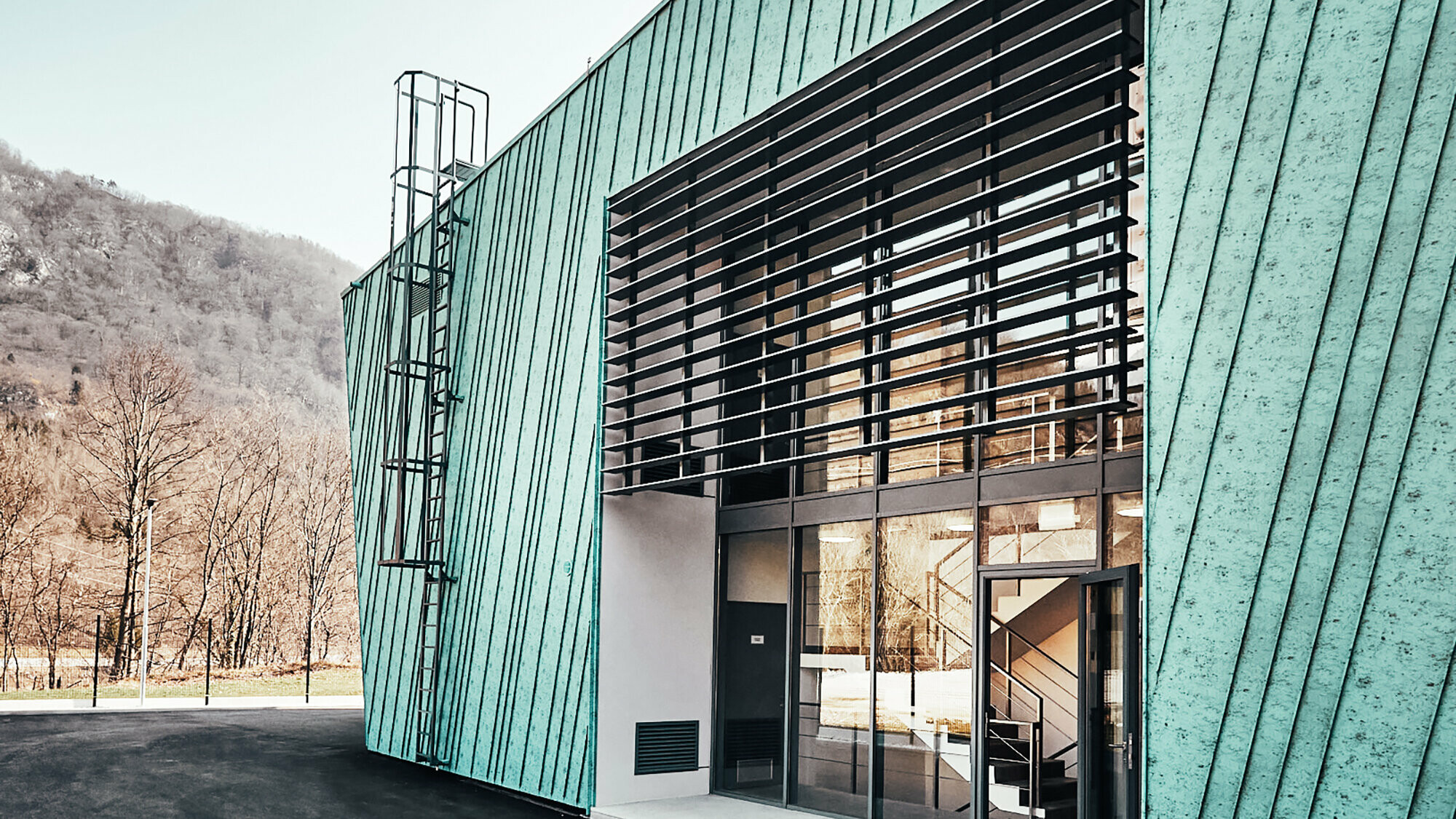 Detail mit leicht schräg verlaufender Fassade mit kleinem, länglichem Fenster, in dem sich Berge und elektrische Leitungen widerspiegeln. Seitenansicht auf Fassade und Glasfront mit Eingang.