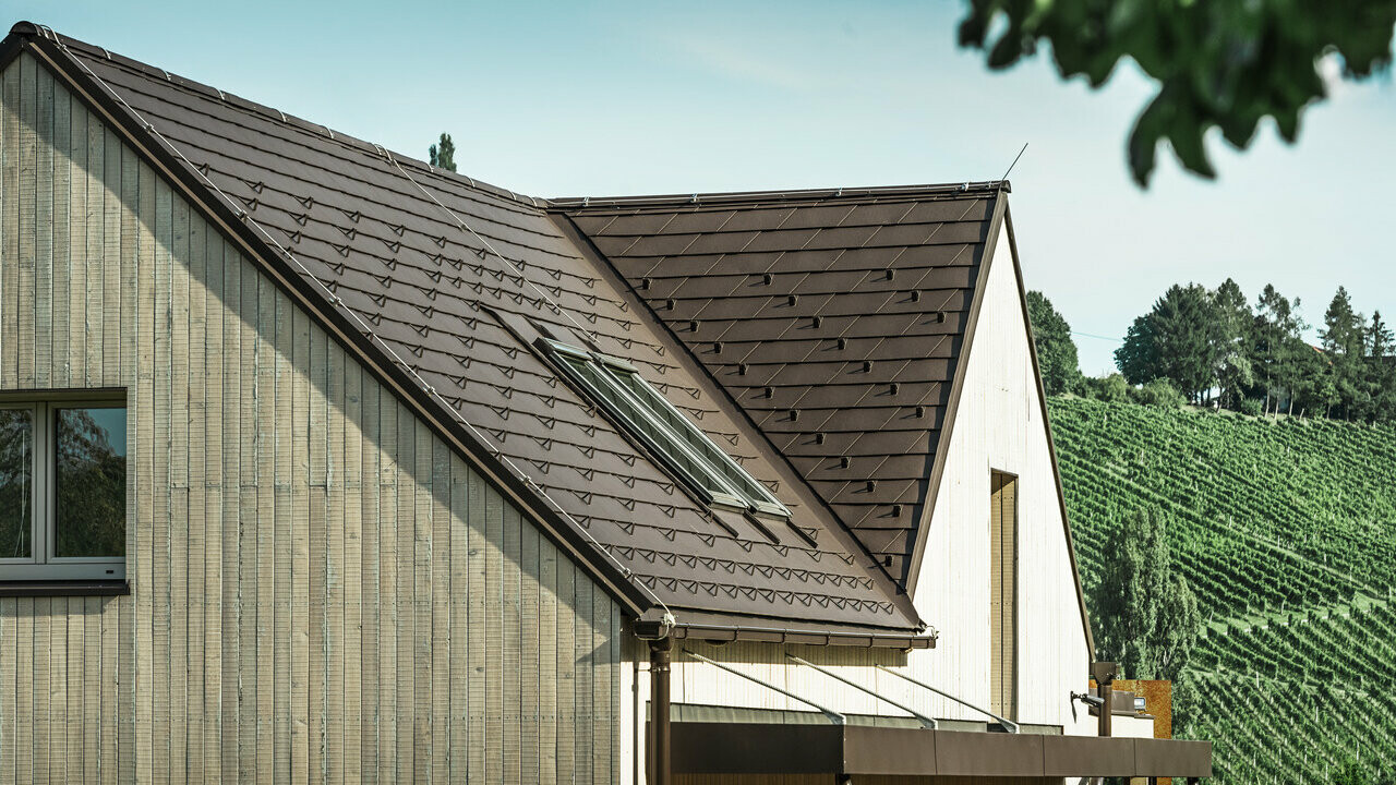 Cette maison individuelle avec deux toits à pignon est recouverte de bardeaux de toiture PREFA en brun noisette. L'écoulement des eaux de pluie se fait par la gouttière carrée PREFA. La façade est revêtue de bois patiné.