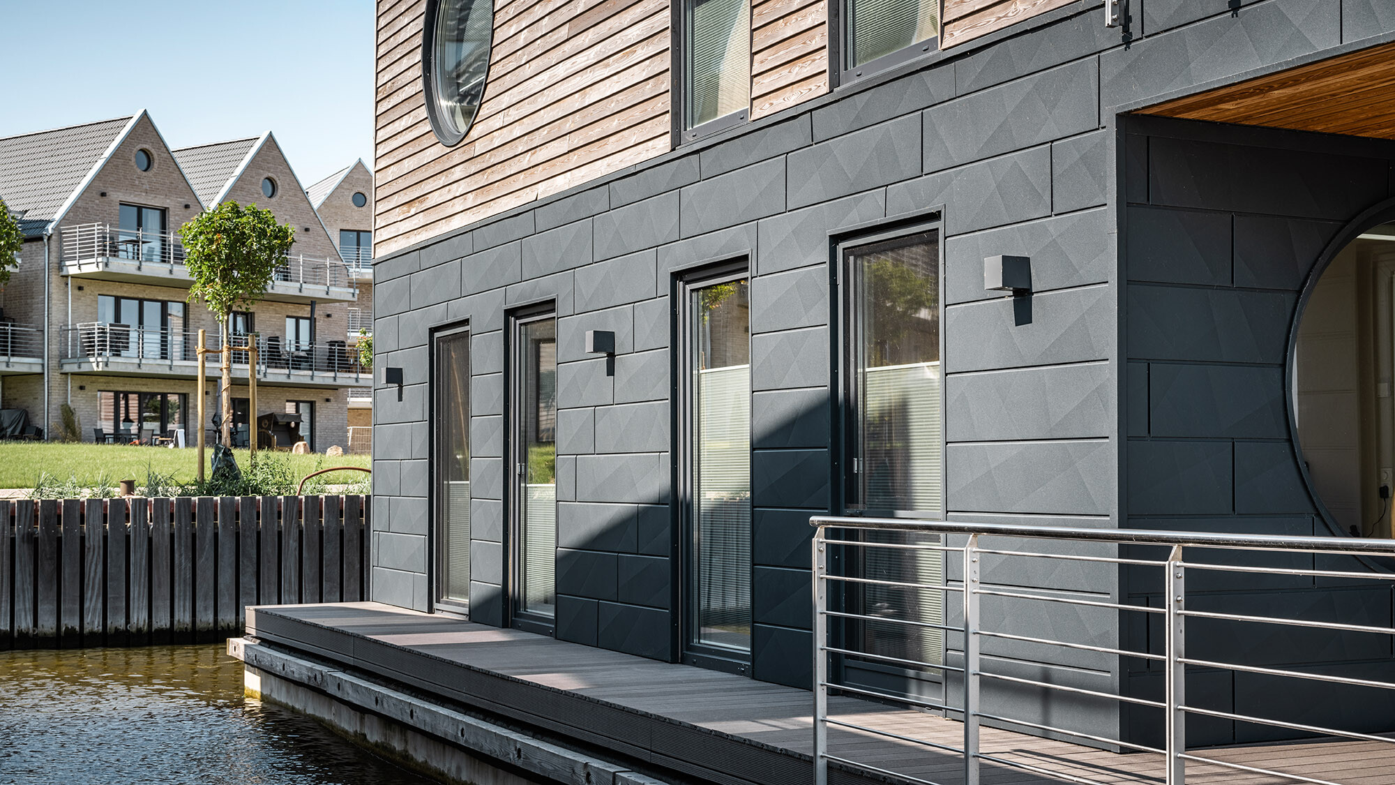 La façade en Siding.X vue de près ; juste devant, on aperçoit la Schlei, tandis qu’une rangée d’immeubles d’habitations construits sur la terre ferme est visible en arrière-plan.