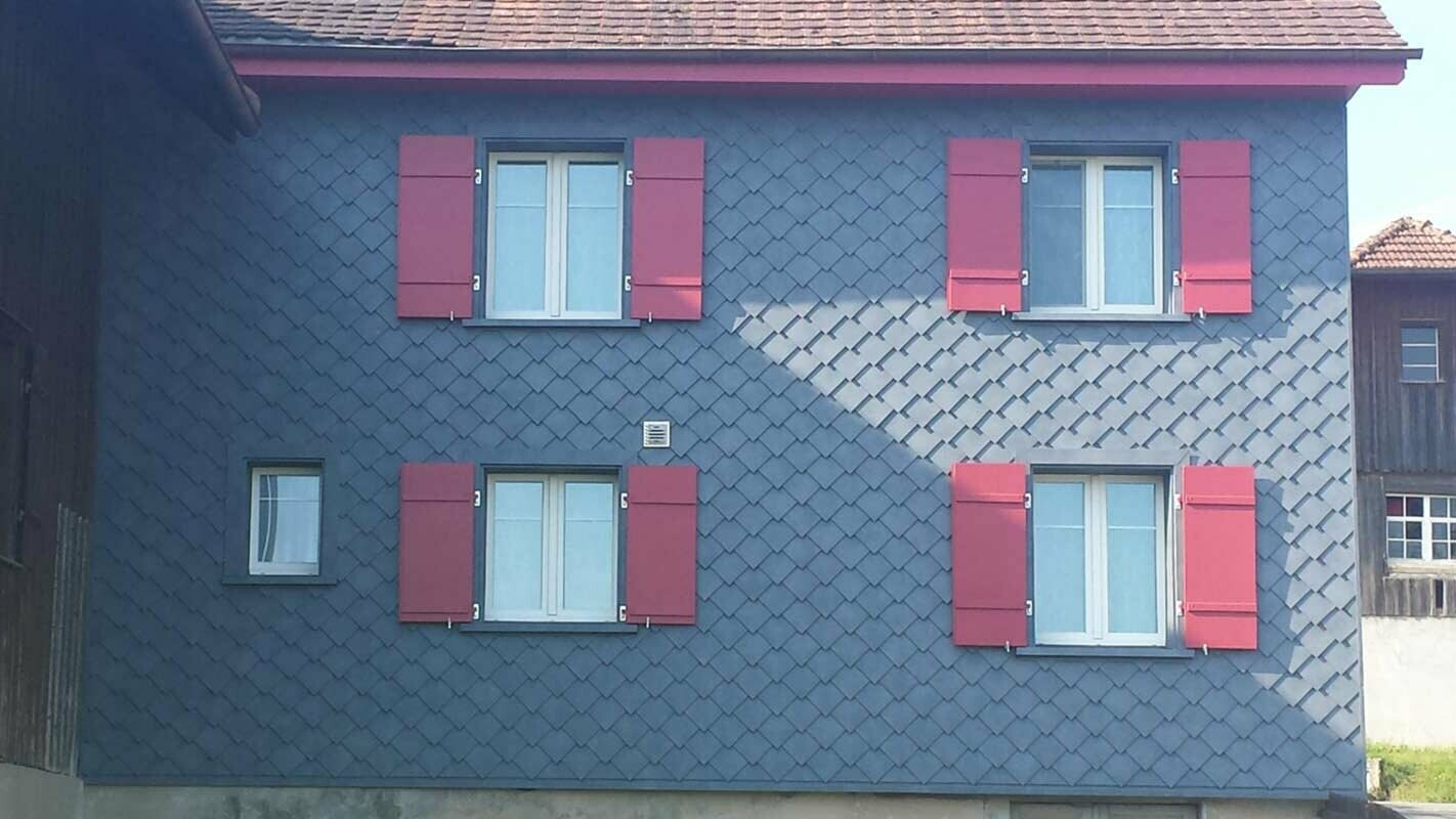 Rénovation de façade à l’aide de losanges de façade PREFA, avec quatre fenêtres dotées de volets rouges