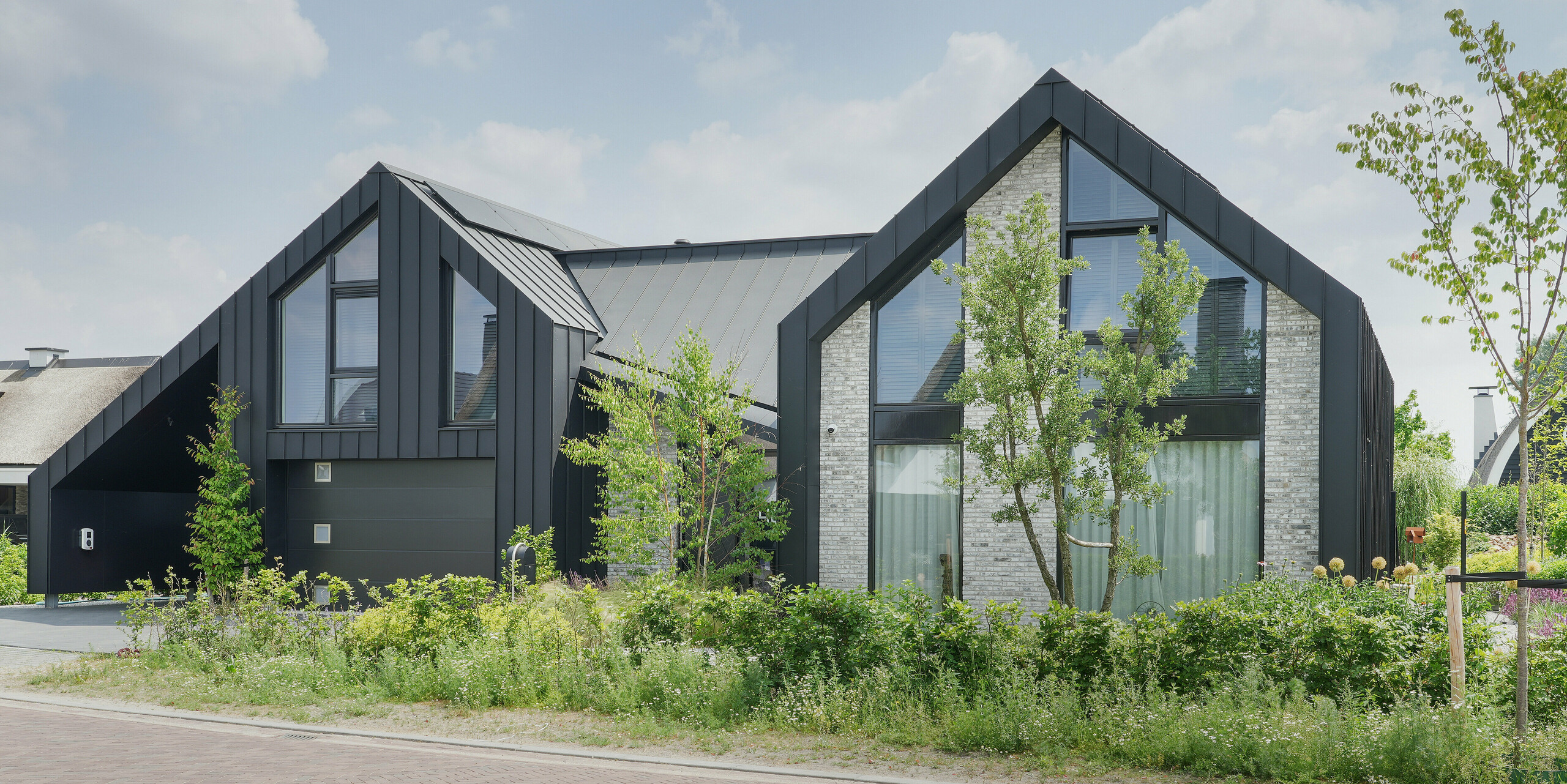 Un aperçu d'une maison individuelle moderne à Veenendaal, aux Pays-Bas, avec un toit noir remarquable et une façade PREFALZ. La maison présente une interaction dynamique de deux structures de pignon reliées par un élément de toit plat. Des accents de pierre naturelle gris clair sur la façade allègent le schéma de couleurs sombres et apportent une structure qui contraste avec les grandes baies vitrées. Le projet combine des lignes claires et modernes avec des éléments naturels afin de créer un lien entre l'espace de vie et le paysage environnant.