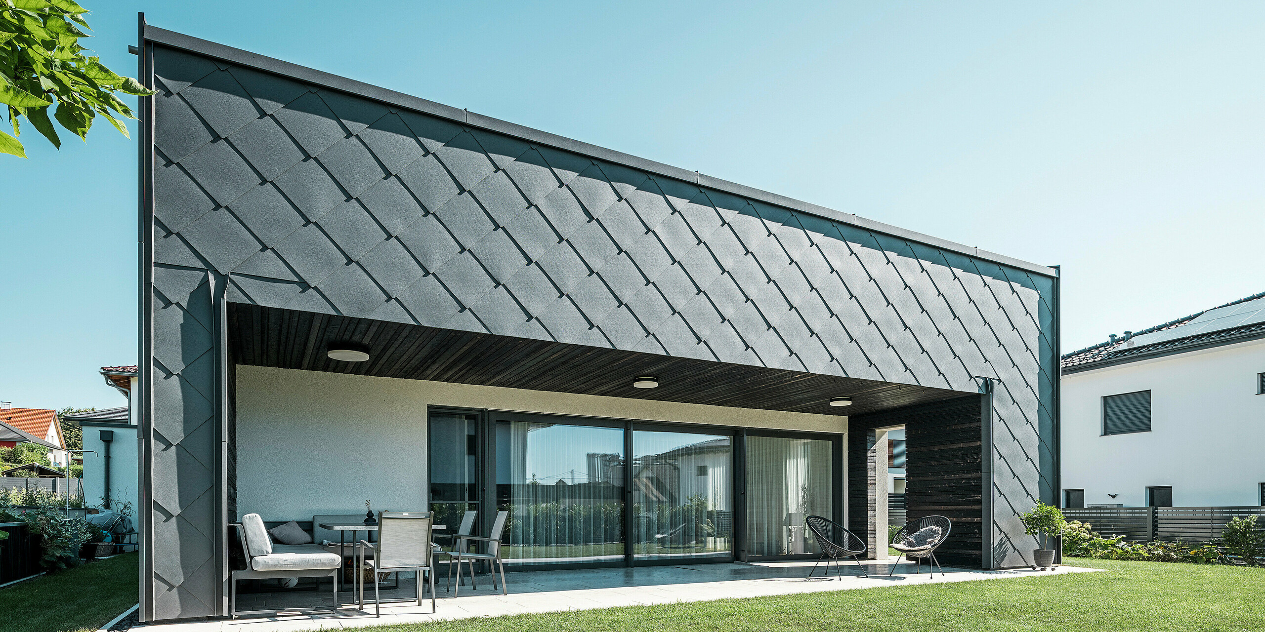 Façade moderne d'une maison unifamiliale à Aschach, revêtue de losanges de façade 44x44 P.10 gris sombre. La terrasse spacieuse et couverte offre un espace extérieur confortable avec des sièges, idéal pour se détendre. Le revêtement en aluminium offre un aspect élégant et une protection optimale contre les intempéries, tandis que les généreuses façades en verre donnent à la maison un aspect lumineux et ouvert.