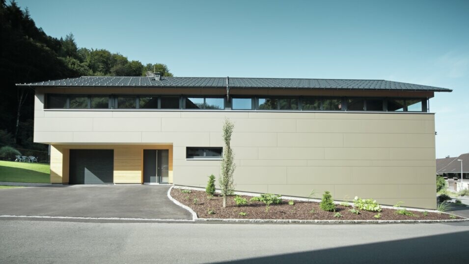 Das Einfamilienhaus in Hohenems präsentiert sich in einer modernen und stilvollen Fassade aus PREFA Aluminium Verbundplatten in Bronzemetallic, mit einem darüberliegenden Dach aus PREFA Dachplatten in P.10 Anthrazit. Die klare Linienführung und das durchdachte Farbkonzept des Designs integrieren das Gebäude nahtlos in die natürliche Umgebung. Die Gestaltung zeigt die Kombination aus Funktionalität und Ästhetik, die PREFA Produkte für zeitgemäße Wohnarchitektur bieten.