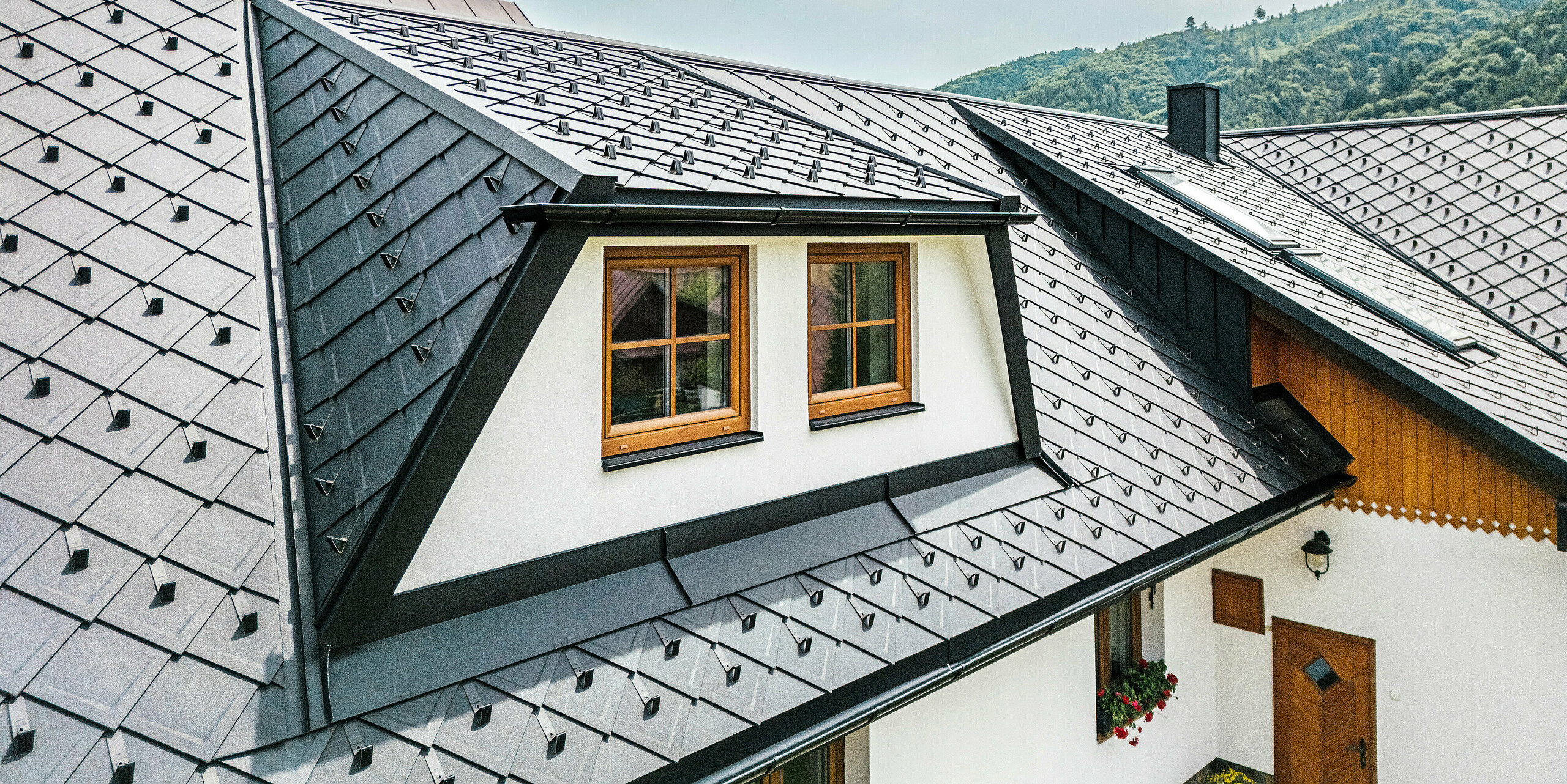 Gros plan du toit d'une maison familiale traditionnelle à Stara Bystrica, en Slovaquie, avec une lucarne trapézoïdale recouverte d'une toiture en losanges PREFA 29 × 29 en P.10 Noir. La toiture en aluminium durable donne au toit un aspect moderne et élégant. La lucarne parfaitement intégrée et les fenêtres avec revêtement en bois s'intègrent harmonieusement dans le paysage du toit et mettent des accents architecturaux.