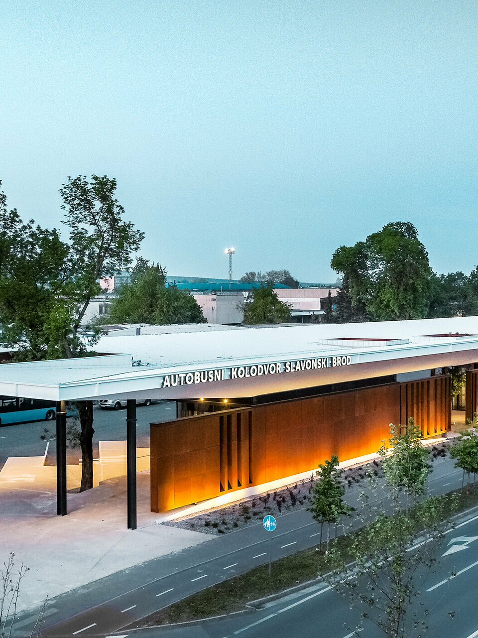 La photo montre le hall de la gare routière « Autobusni Kolodvor Slavonski Brod » en Croatie au crépuscule. Le hall de la gare est doté d'un grand toit blanc Prefalz de PREFA, soutenu par des colonnes noires. La façade se compose de panneaux bruns modernes, éclairés par des lampadaires, créant une ambiance chaleureuse et accueillante. Sous le toit, on peut voir les bus et les espaces d'attente. À l'arrière-plan, on distingue divers bâtiments, dont une grande tour de bureaux et plusieurs immeubles résidentiels.