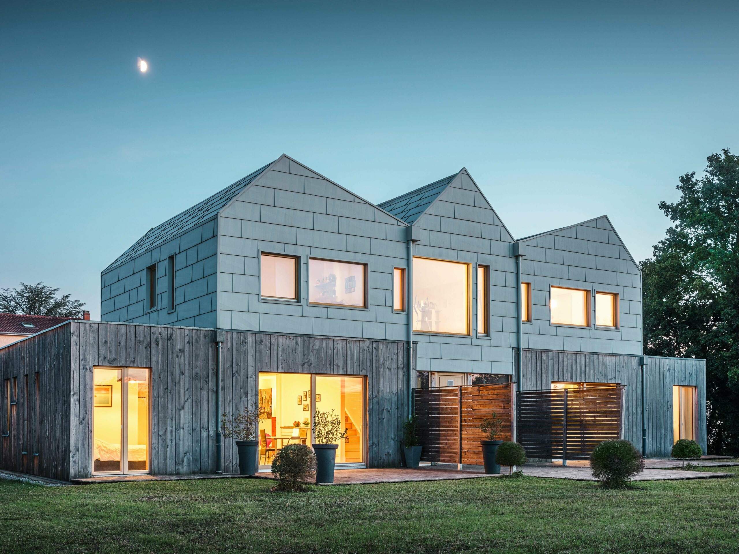 Maison jumelée au crépuscule avec des intérieurs lumineux visibles à travers de grandes fenêtres. Les panneaux en aluminium de PREFA au look FX.12 s'intègrent parfaitement dans le design moderne. Le bâtiment est entouré d'un jardin bien entretenu.