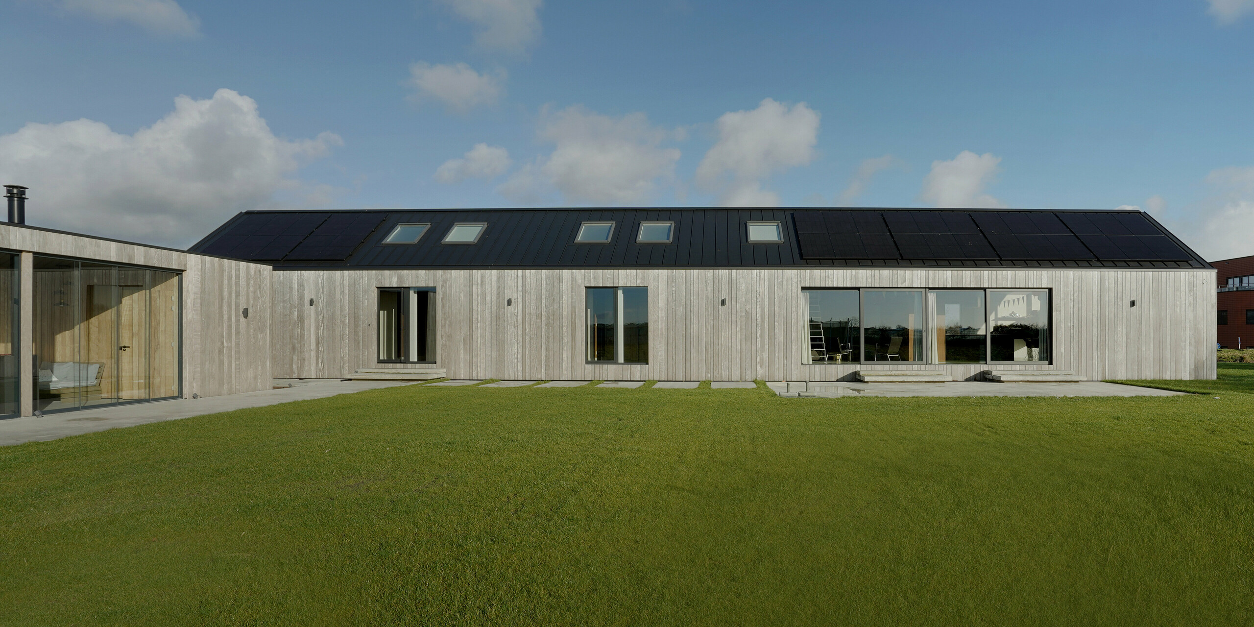 Vue de face de tout le côté large d'une maison familiale moderne à Uithoorn, aux Pays-Bas, avec toit à joints debout PREFALZ en gris noir et système photovoltaïque. La photo montre le bâtiment allongé avec de grandes façades vitrées et un revêtement en bois, entouré d'une pelouse bien entretenue. Les lignes claires et l'utilisation de matériaux de haute qualité résistants aux intempéries comme l'aluminium et le bois durable soulignent le design écologique et moderne de la maison. Idéal pour les projets de construction respectueux de l’environnement qui mettent l’accent sur la qualité et l’esthétique.