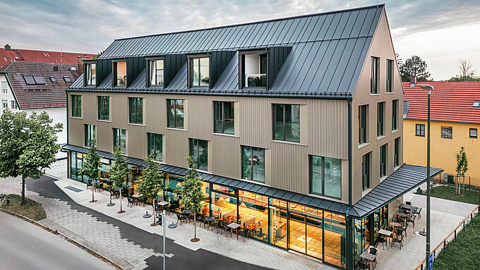 La photo montre un bâtiment moderne avec un revêtement de façade en Prefalz de couleur bronze et un toit à deux versants, également recouvert de Prefalz de couleur gris noir. La grande façade vitrée du rez-de-chaussée est encadrée d'arbres et crée une atmosphère invitante pour le restaurant situé en contrebas. La combinaison des matériaux Prefalz de haute qualité et de l'architecture moderne confère au bâtiment un aspect élégant et contemporain.