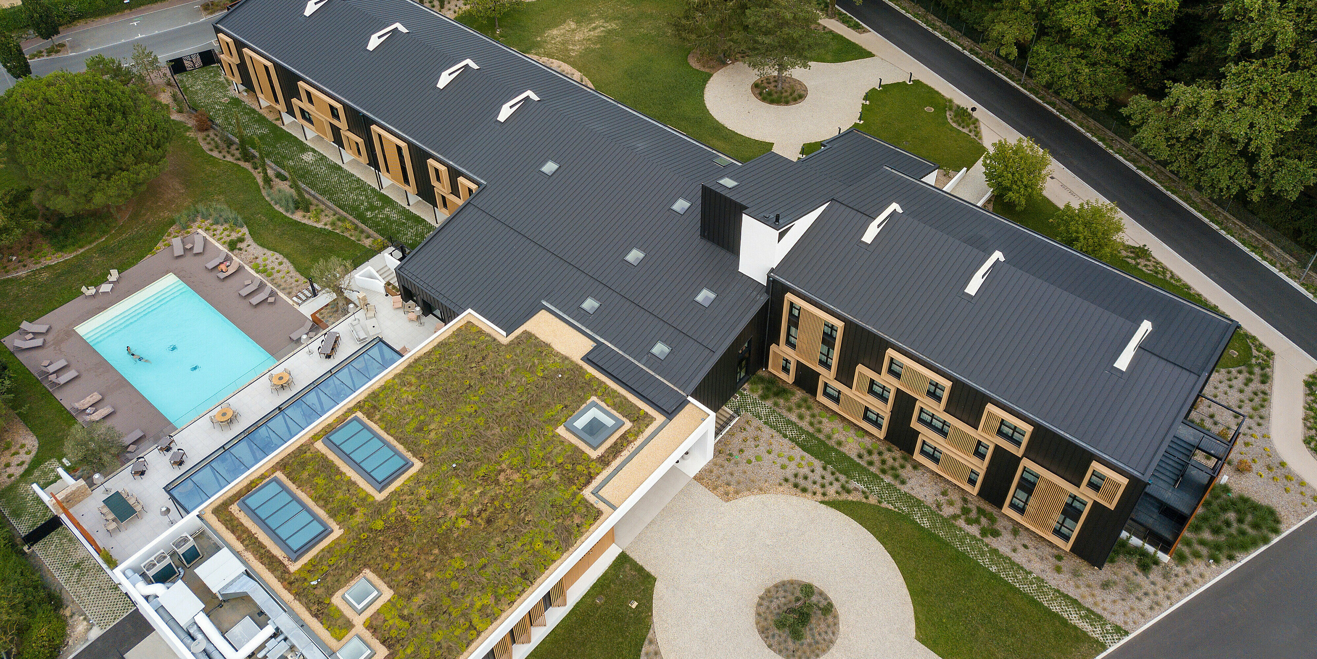 Vue aérienne de la zone d'entrée de l'Hôtel Parc du Landreau aux Herbiers, France. Le bâtiment a été revêtu du système de toiture et de façade PREFALZ en gris noir. Les cadres en bois frappants autour des fenêtres complètent le revêtement moderne en aluminium. L'espace extérieur avec piscine et toit partiellement végétalisé reflète la construction durable de l'hôtel.