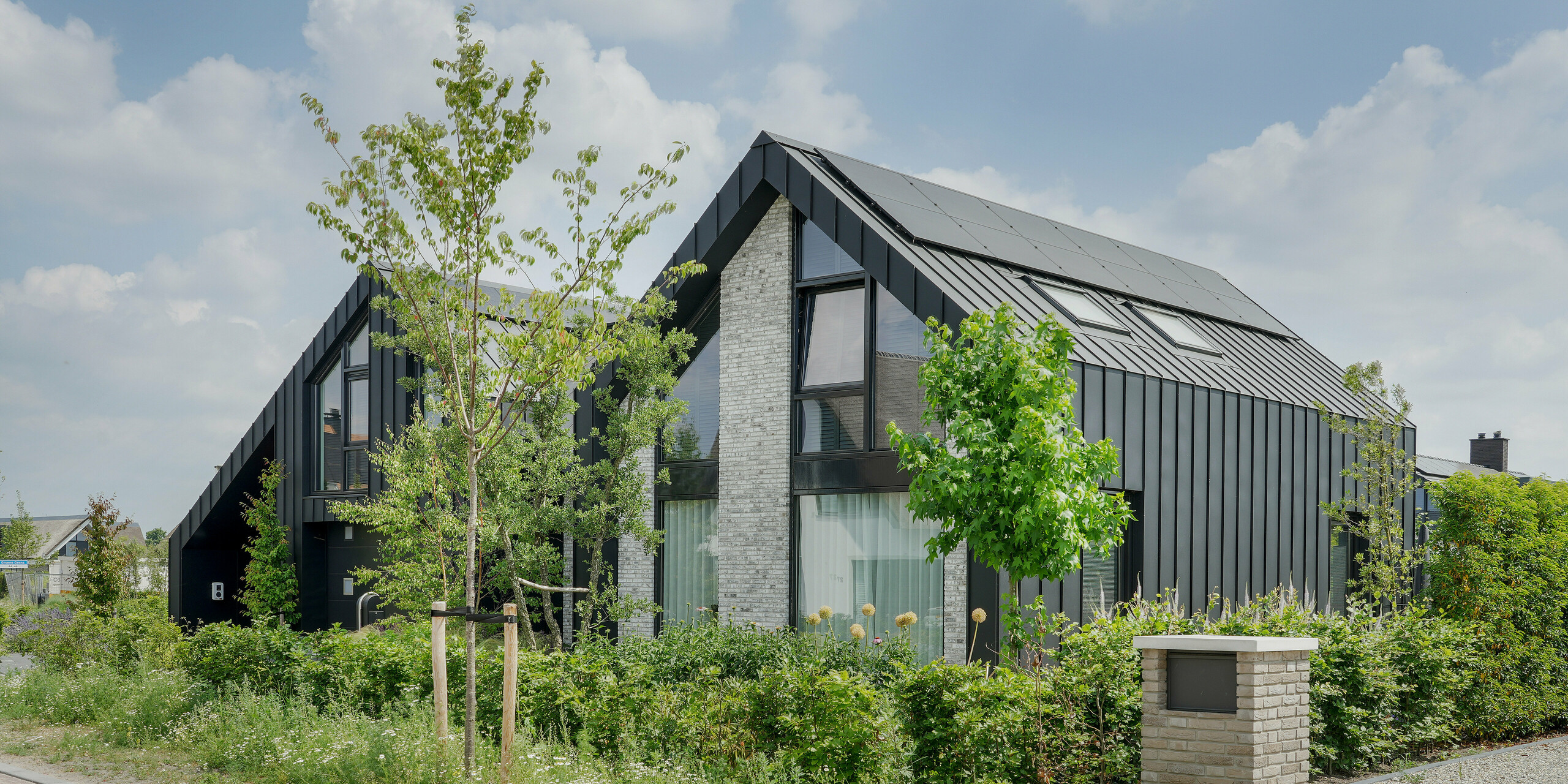 Architecture résidentielle moderne à Veenendaal, Pays-Bas, caractérisée par l'aluminium noir de PREFA sur le toit et certaines parties de la façade. La maison, avec ses pignons pointus et son toit en gradins, a une forme géométrique très marquée. L'utilisation de pierre naturelle gris clair comme partie de la façade offre un contraste subtil avec les tuiles à joint debout foncées. La maison est entourée d'un jardin naturel et sauvage et d'allées pavées qui créent un lien entre l'architecture moderne et l'environnement naturel.