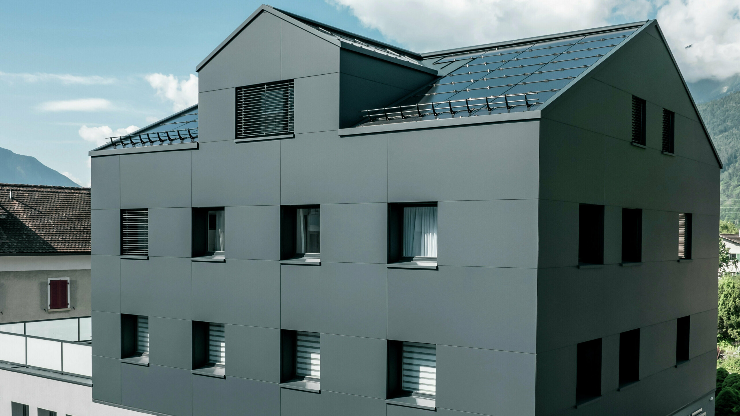 Modernisiertes Wohn- und Geschäftsgebäude nach der Fassadensanierung mit PREFABOND Aluminium Verbundplatten in Anthrazit. Die klare, elegante Linienführung der neuen Fassade verleiht dem Gebäude ein modernes Erscheinungsbild. Im Erdgeschoss befindet sich das Architekturbüro Voser. Die Umgebung mit Bäumen und Bergen rundet die harmonische Optik ab.