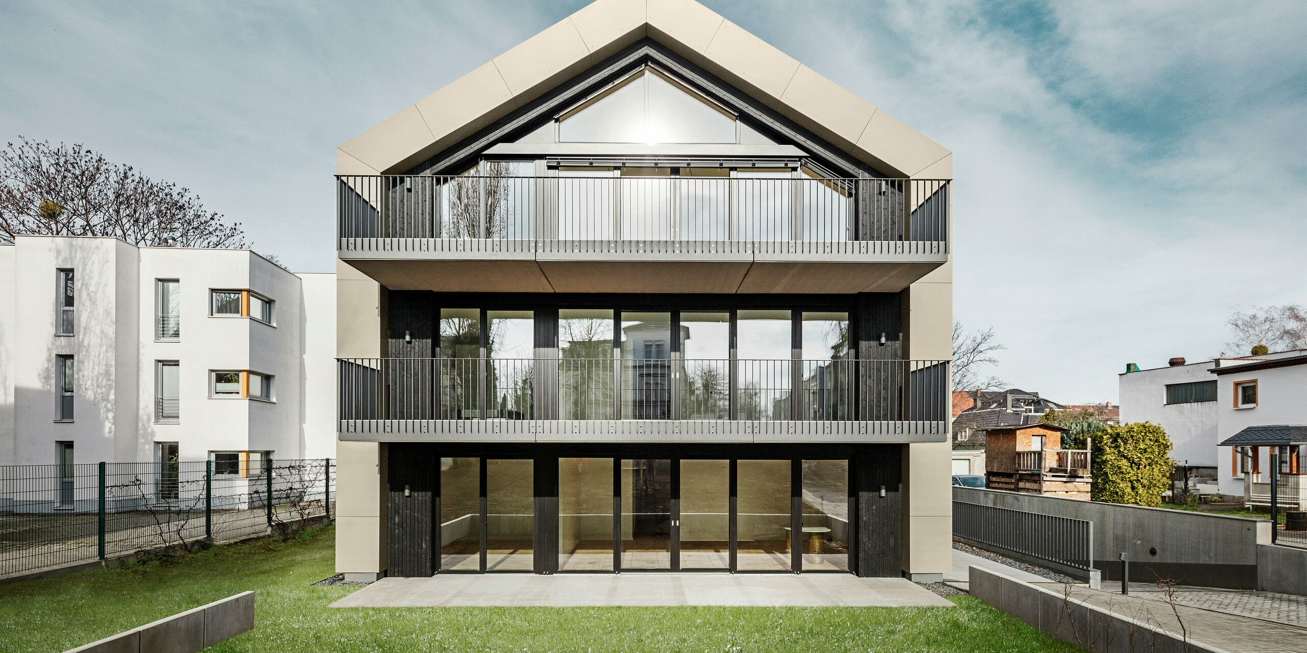 Vue de face d'un immeuble d'appartements moderne à Dresde (Chemnitzer Straße) avec une impressionnante façade en verre et des balcons. La façade et le toit du bâtiment sont revêtus du losange en aluminium durable PREFA 29 × 29 en bronze, qui donne au bâtiment un aspect élégant et contemporain. La photo montre le design symétrique de la façade, les surfaces vitrées généreuses et le jardin bien entretenu au premier plan.