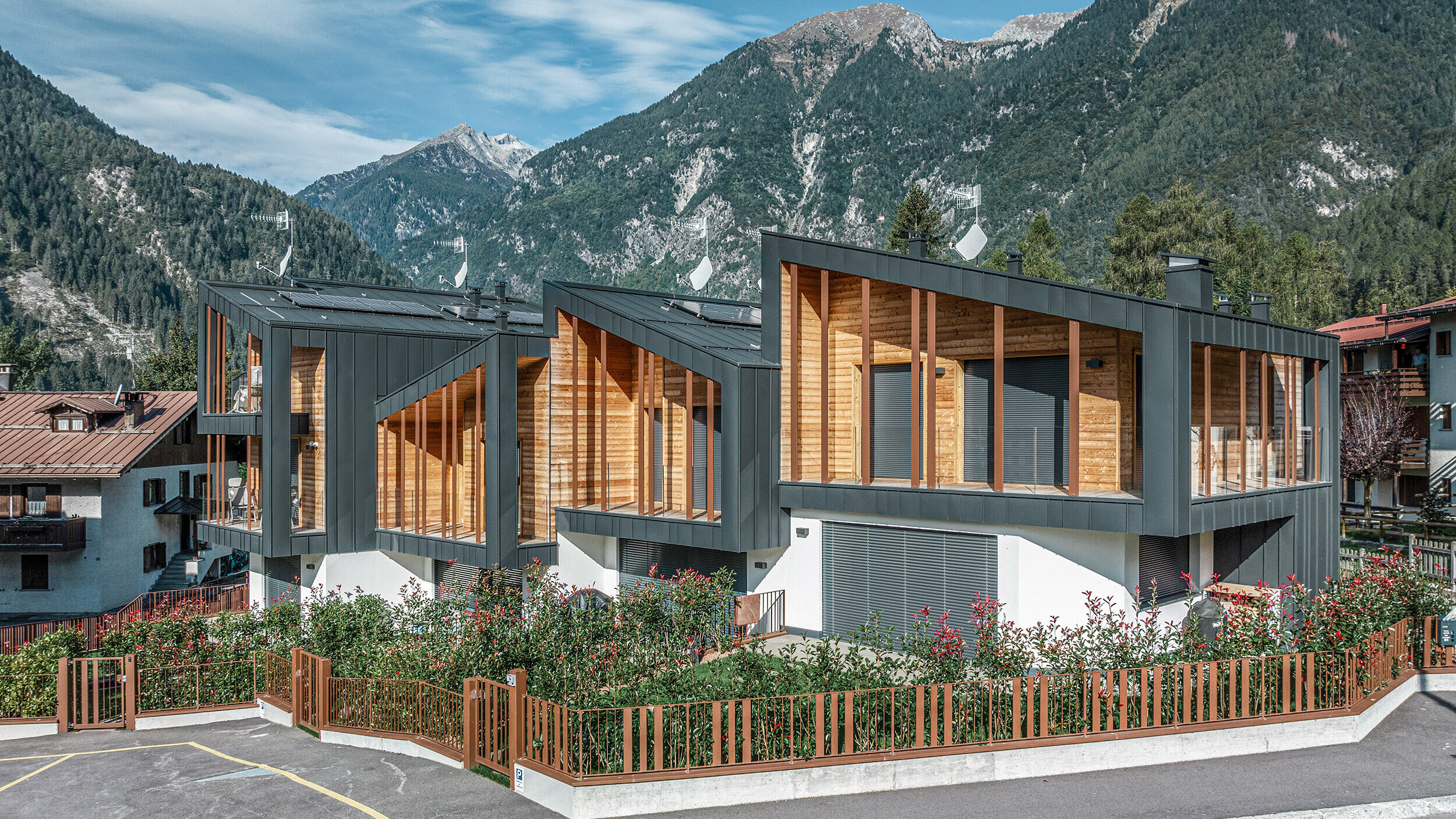 Nouvelle construction de chalets modernes avec toits monopentes en métal Prefalz, façades associant métal gris foncé et éléments en bois, le tout dans un paysage de montagne et sous un ciel dégagé. Les larges fenêtres et terrasses sont complétées par des espaces verts et un parking entouré d’une clôture.