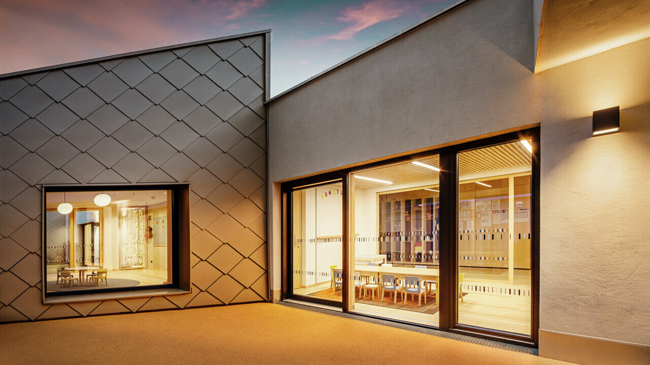 Vue du soir d'une partie du centre de formation de Gerlos, dont la façade a été conçue de manière artistique. À gauche, on peut voir une fenêtre entourée d'un cadre mural PREFA 44x44, tandis qu'à droite, l'entrée attire l'attention avec une généreuse baie vitrée qui donne sur un intérieur bien éclairé avec des chaises et des tables. L'éclairage intérieur chaleureux contraste avec la lumière tamisée du crépuscule, ce qui met en valeur l'architecture moderne et le design de la façade. 