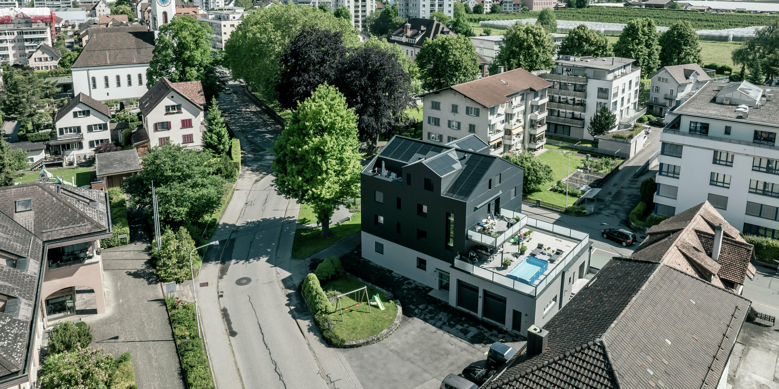 Vue d'un bâtiment commercial moderne avec une façade rénovée en panneaux composites en aluminium PREFABOND en anthracite. Le bâtiment s'intègre harmonieusement dans l'environnement verdoyant et les immeubles résidentiels environnants, tandis qu'un impressionnant paysage de montagne peut être vu en arrière-plan.