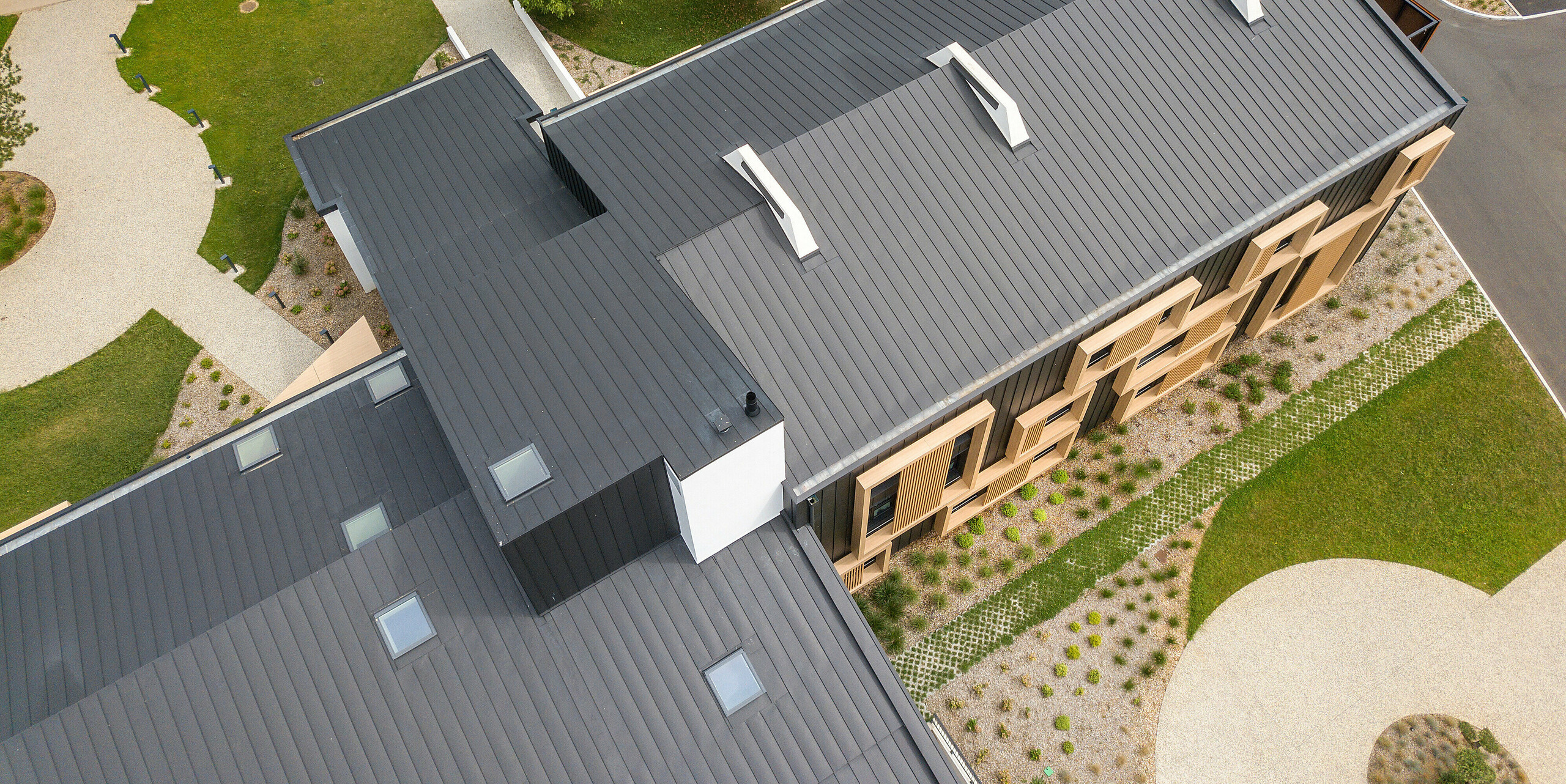 Vue aérienne de l'Hôtel Parc du Landreau aux Herbiers, France, avec une couverture de toit à joint debout uniforme sous forme de PREFALZ en gris noir. Le système de toiture moderne en aluminium impressionne par sa durabilité et sa résistance aux intempéries et s'intègre harmonieusement dans le paysage environnant. Les lignes claires du toit à joint debout complètent parfaitement la conception architecturale du bâtiment.