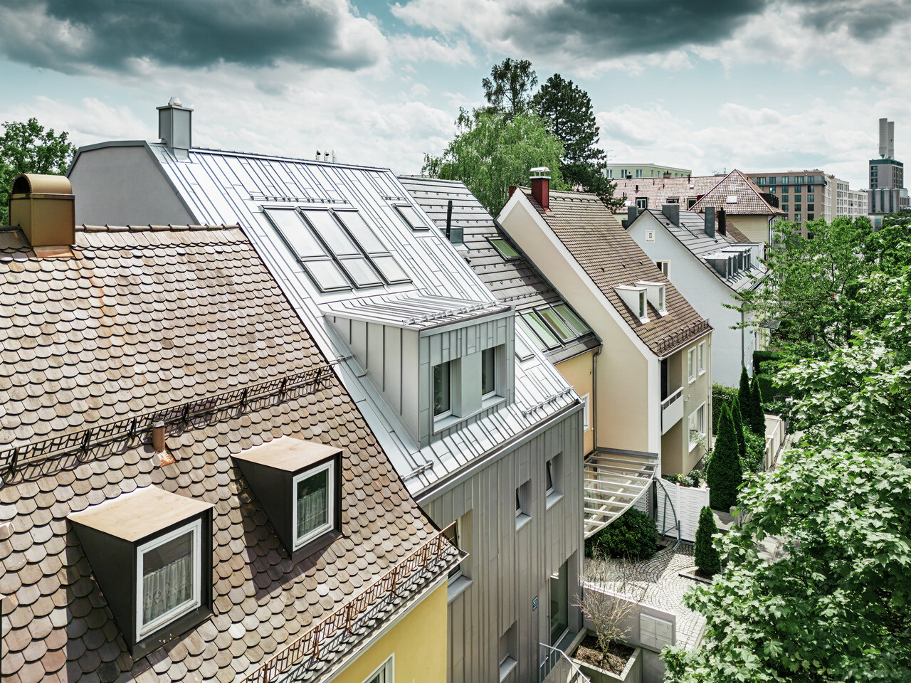 Vue sur les toits d'un lotissement de maisons mitoyennes avec un mélange de toits en tuiles traditionnels et de toits modernes en aluminium Prefalz métallisé argenté. Une maison se distingue avec son toit rénové aux reflets métalliques et ses lucarnes assorties. La végétation verte et dense des jardins complète l'image et crée un contraste naturel avec l'architecture urbaine. En arrière-plan, on distingue les contours de constructions urbaines modernes.