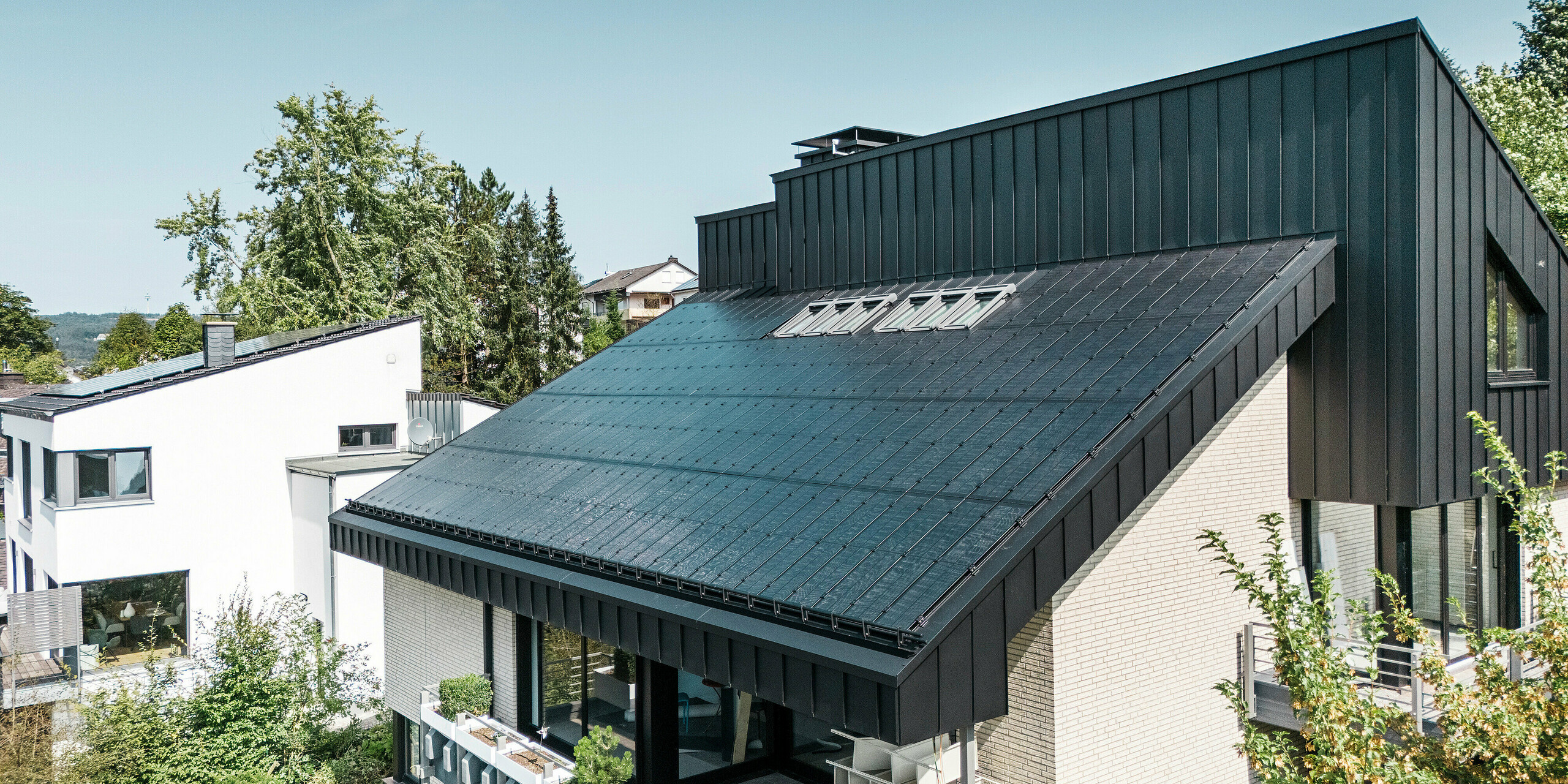 Architecture moderne d'une maison unifamiliale à Meschede, en Allemagne, avec une toiture en aluminium PREFALZ en P.10 noir et des modules photovoltaïques verre/verre intégrés. Le toit en pente décalé allie un design intemporel à une technologie photovoltaïque durable. Les modules solaires innovants PREFALZ délivrent une puissance de 18 kWc. Les modules sans cadre et l'élégant revêtement en aluminium de la façade et du toit forment une unité esthétique et fonctionnelle qui s'intègre parfaitement dans l'environnement de vie vert.